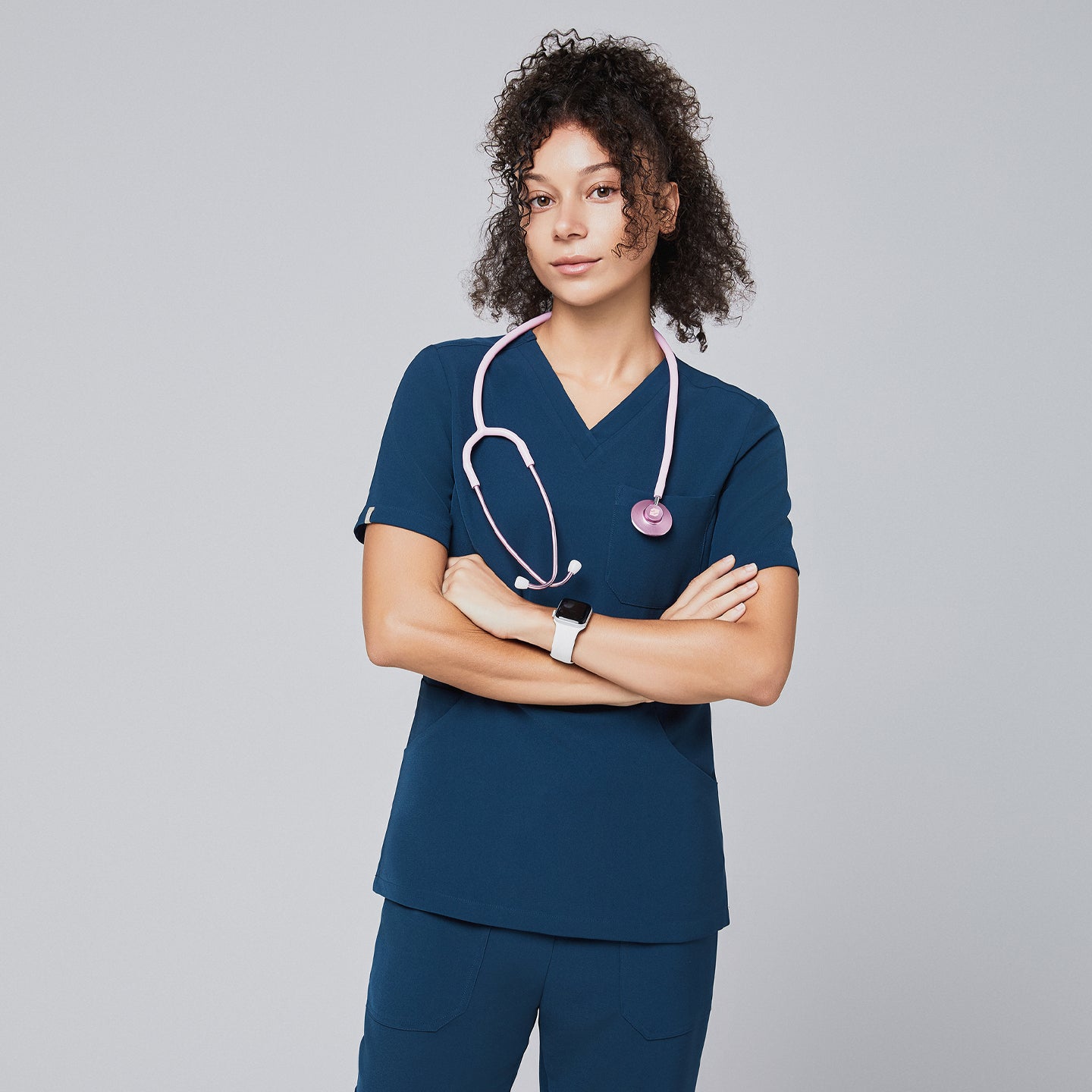 Woman wearing a dark blue V-neck scrub top with a pink stethoscope around her neck, standing confidently with arms crossed, against a light gray background,Dark Blue