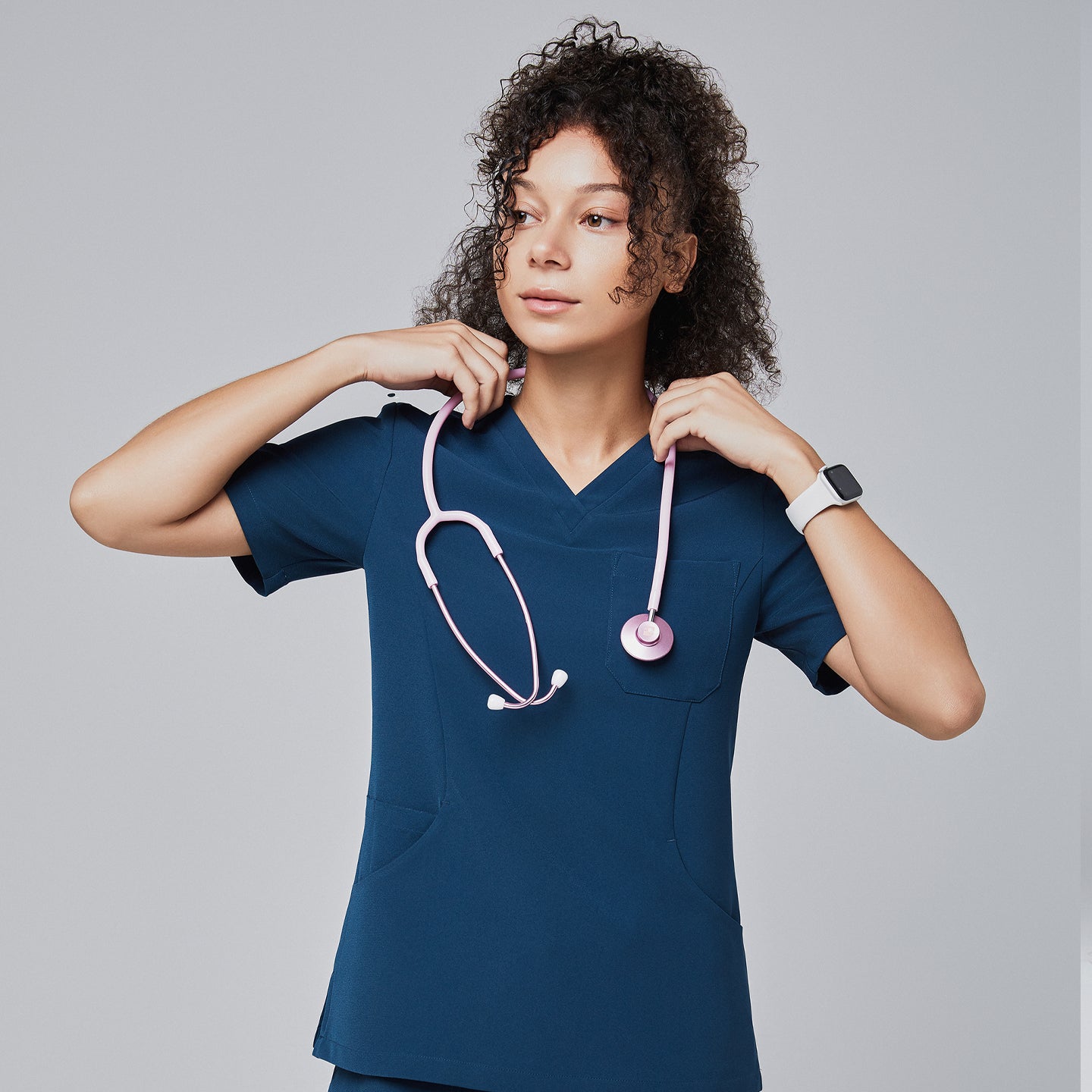 Image of Model wearing a dark blue V-neck scrub top with a pink stethoscope around her neck, looking to the side, standing against a light gray background,Dark Blue