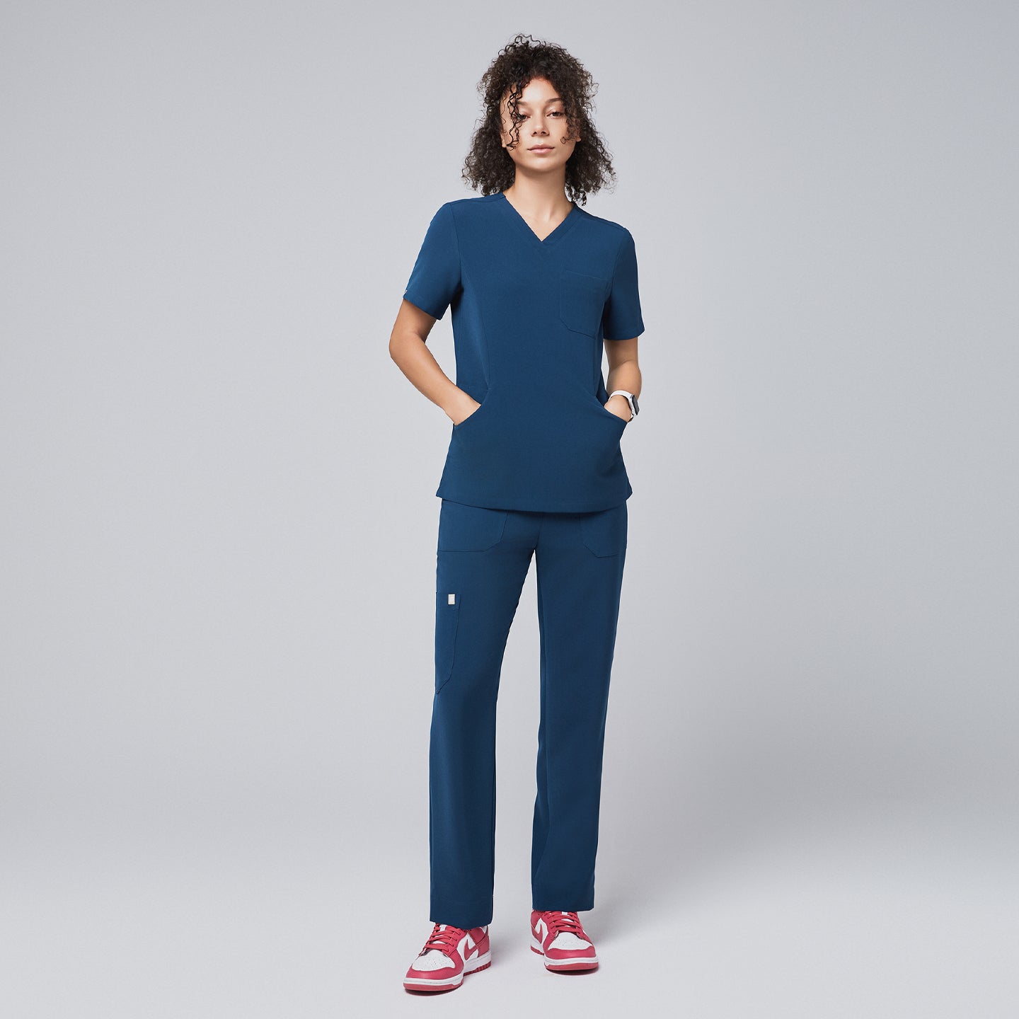 Image of Model dressed in a dark blue V-neck scrub top and matching straight-leg pants with hands in pockets, paired with red and white sneakers, standing against a light gray background,Dark Blue