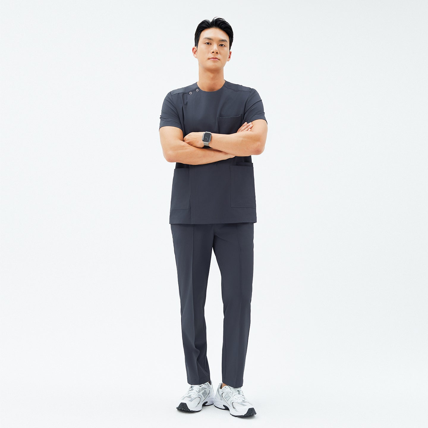 Male model wearing TUL Men’s Premium Banding Pants in Deep Gray, standing with arms folded. The pants have crisp front seams, and the matching scrub top features a snap detail on the shoulder. He pairs the outfit with white athletic shoes.