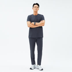 Image of Male model wearing TUL Men’s Premium Banding Pants in Deep Gray, standing with arms folded. The pants have crisp front seams, and the matching scrub top features a snap detail on the shoulder. He pairs the outfit with white athletic shoes.