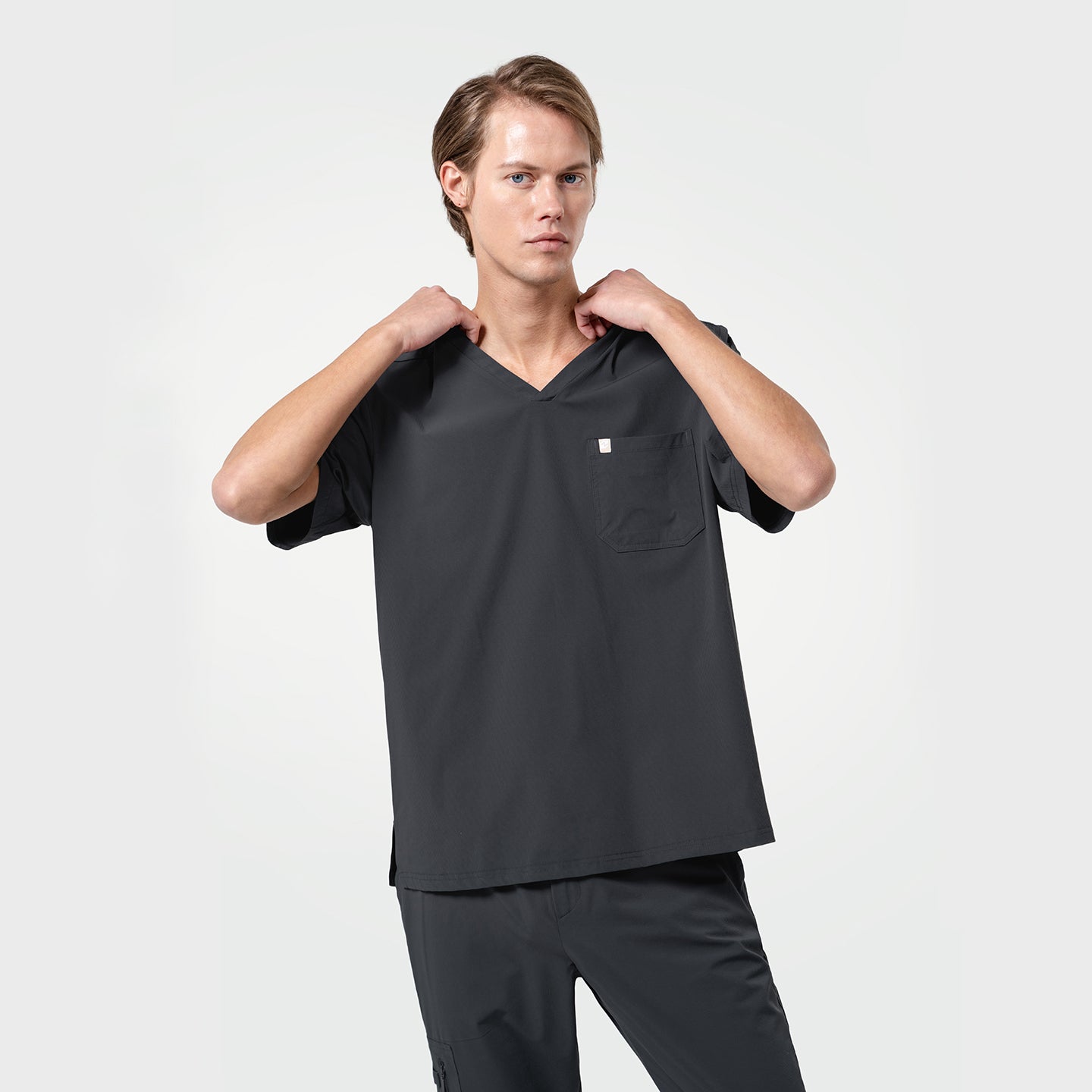 Image of Adjusting the neckline of his scrub top, the male model wears a Deep Gray scrub set, presenting a modern and professional look.