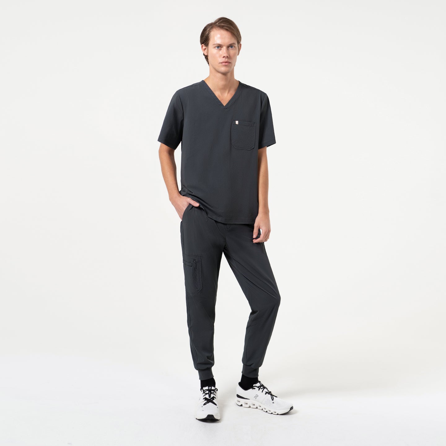 Image of Full-body view of a model wearing a Deep Gray single-pocket scrub top with matching jogger scrub pants, highlighting the relaxed V-neck design and modern, minimal silhouette.