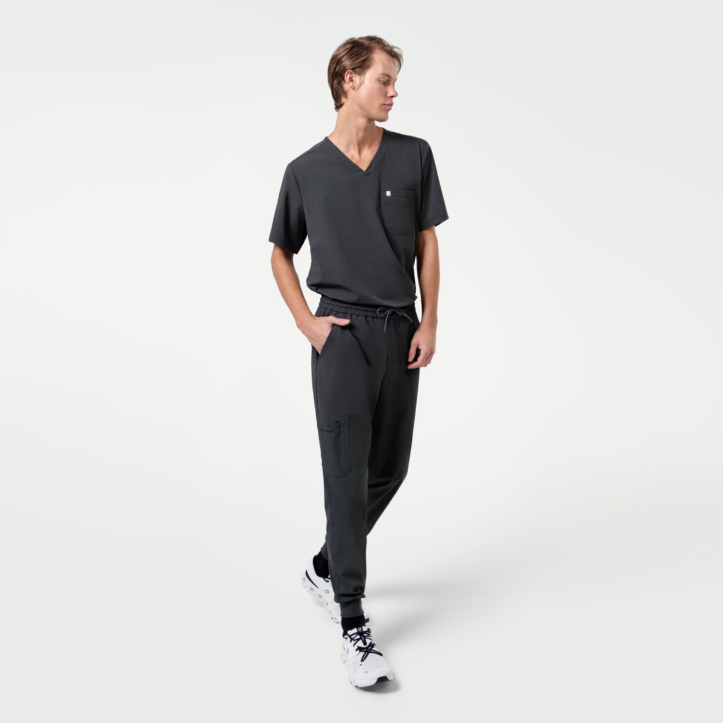 Image of The model wearing the Deep Gray Single Pocket Scrub Top walks casually with one hand in the pocket, looking to the side.