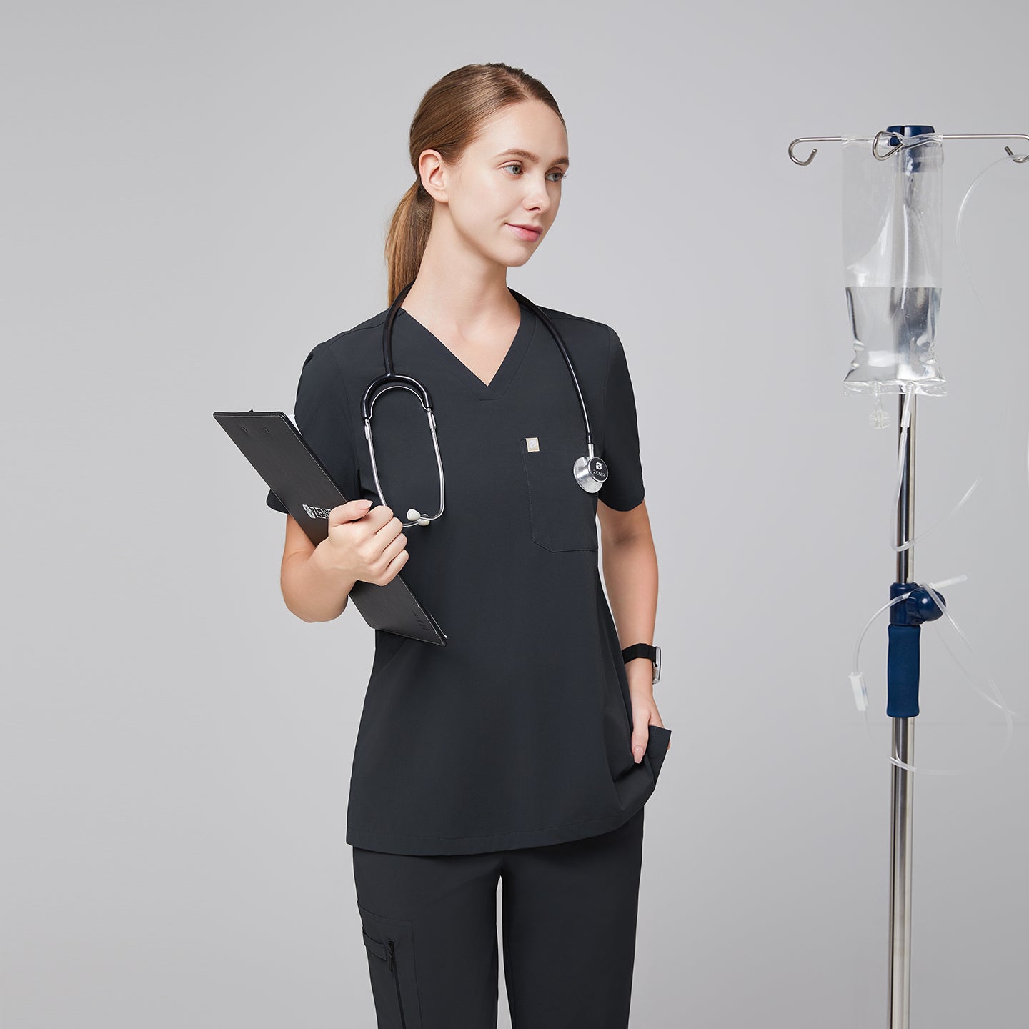 Image of Female healthcare professional wearing a dark gray V-neck scrub top and pants, holding a clipboard with an IV stand in the background,Deep Gray