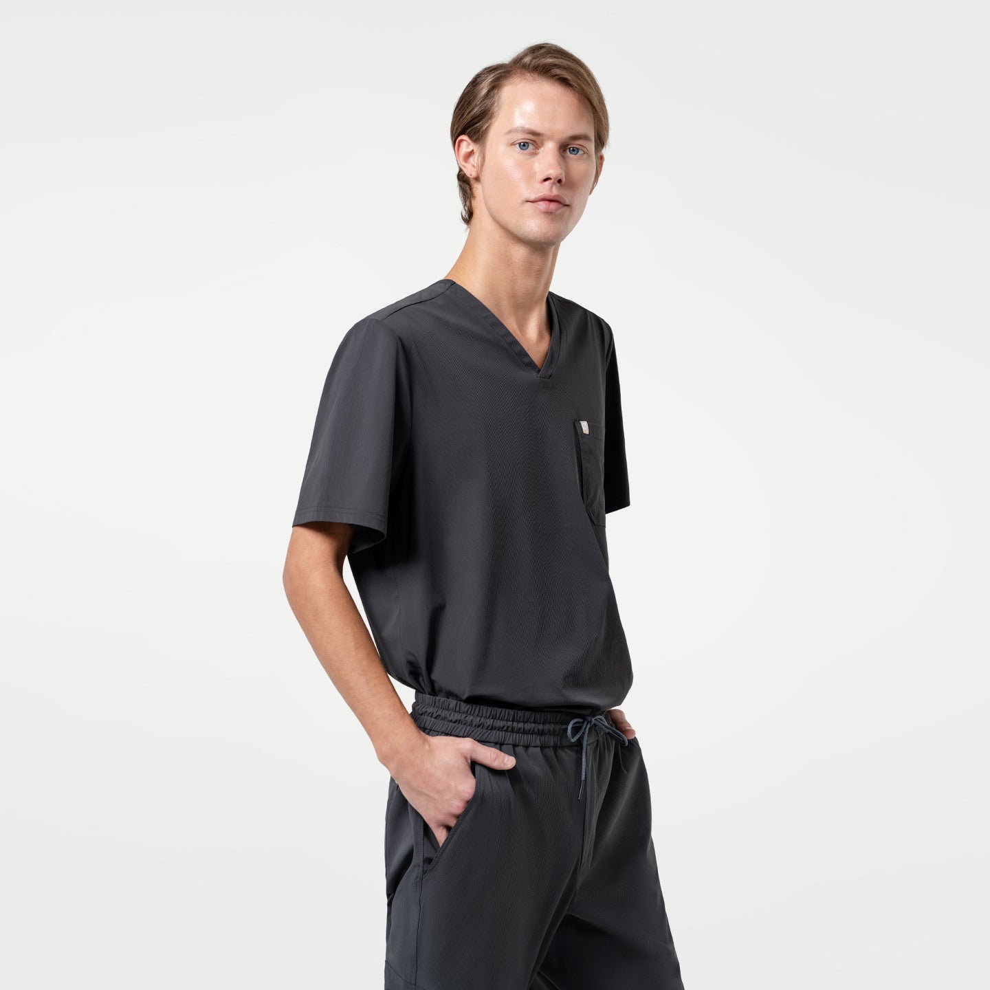 Image of With one hand in his pocket, the male model stands in a Deep Gray scrub set, giving a sleek and professional appearance.