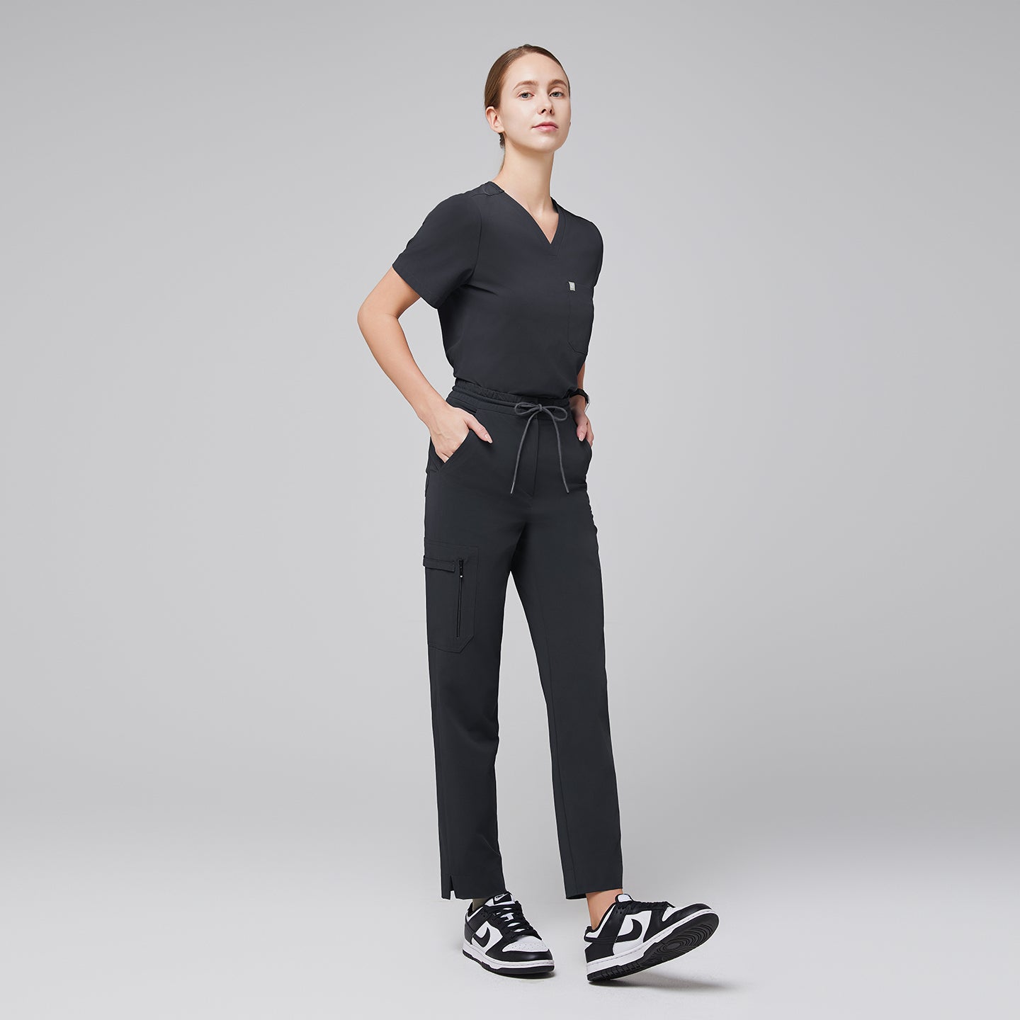 Image of Model wearing a scrub set with a V-neck top and drawstring pants featuring zipper pockets and a split hem, paired with sneakers for a modern professional look,Deep Gray