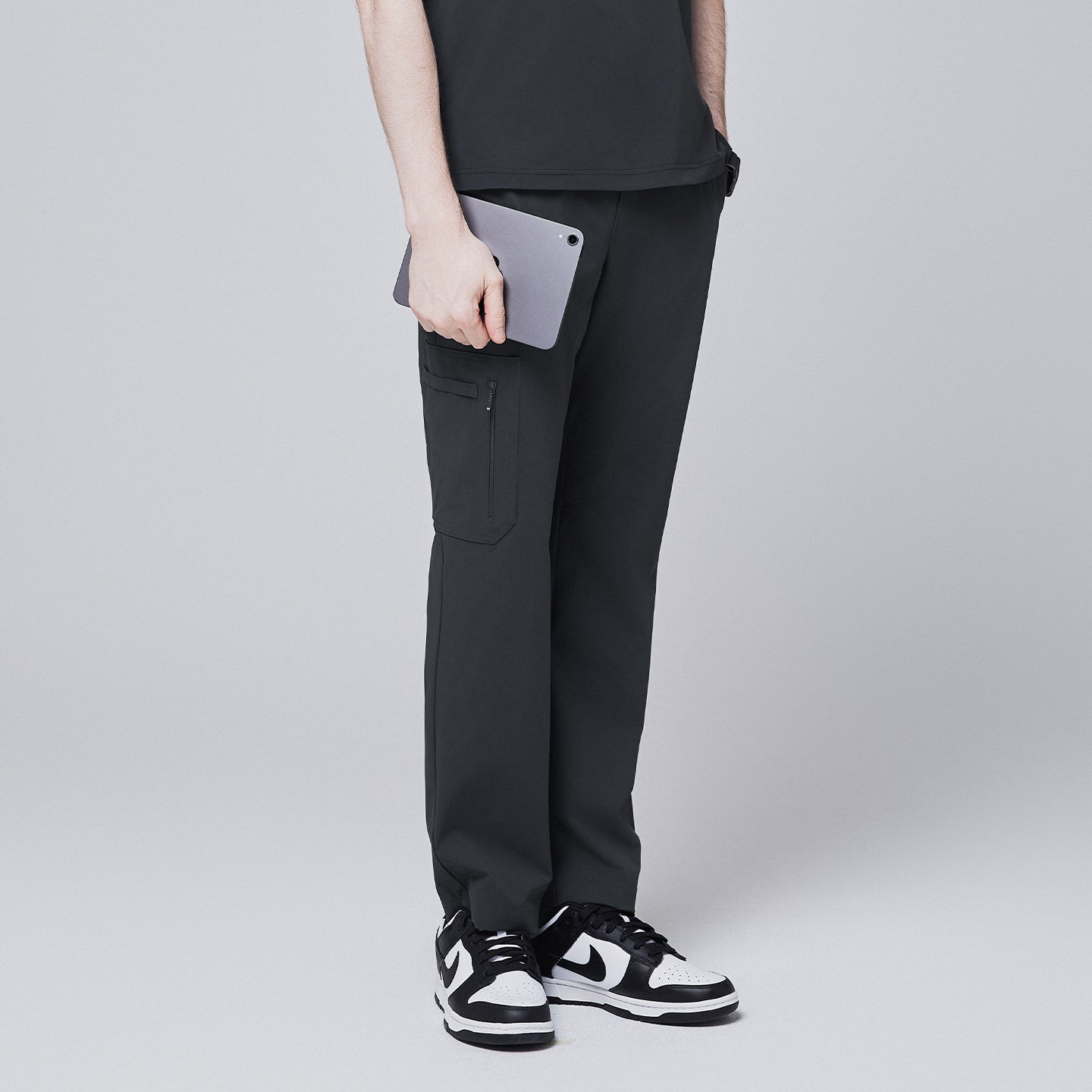 Image of Side view of deep gray straight scrub pants featuring a secure zipper pocket, modeled while holding a tablet, paired with black and white sneakers against a neutral gray background,Deep Gray