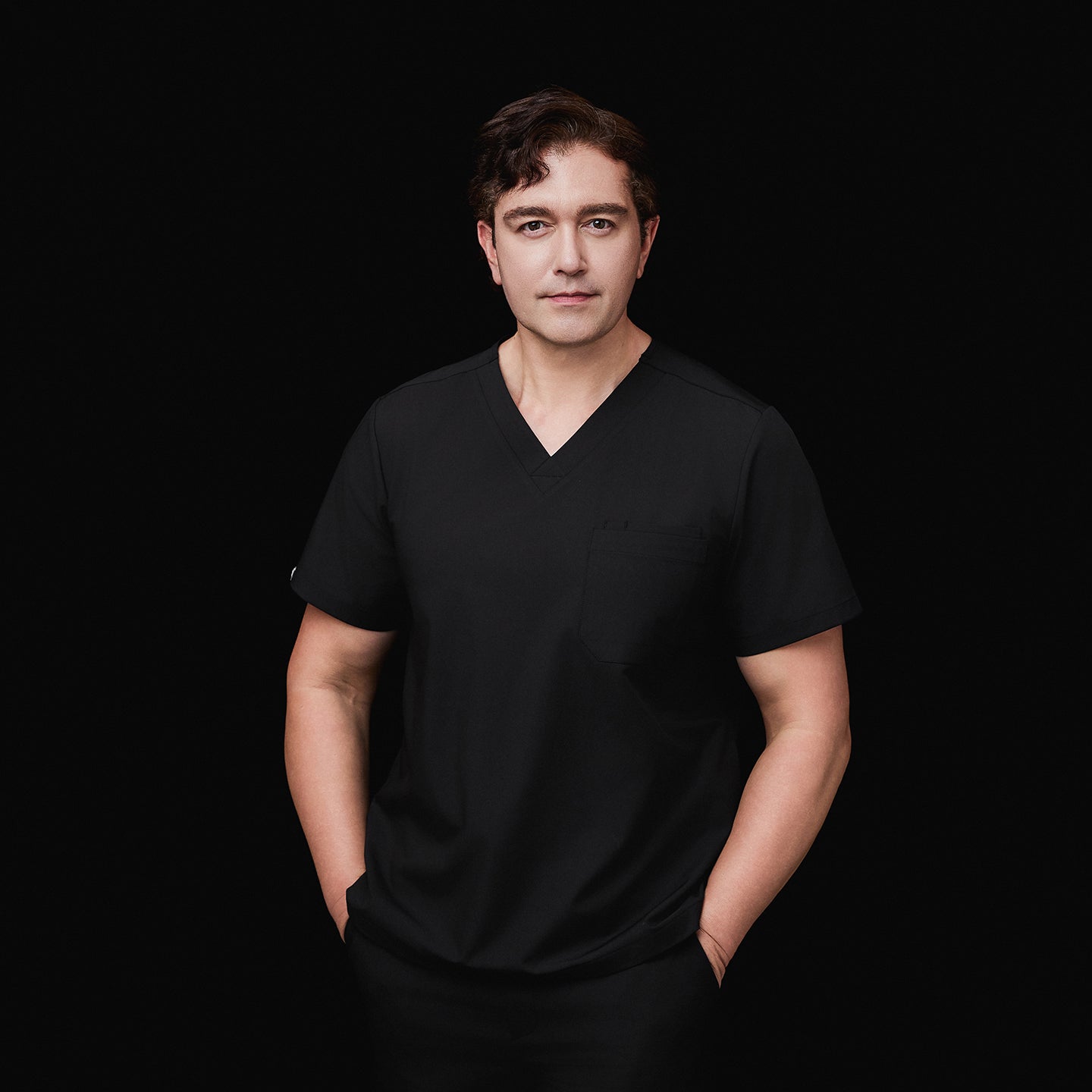 Image of Model wearing an eco black 3-pocket scrub top with a V-neck design, hands in pockets, styled with matching scrub pants, against a black background,Eco Black