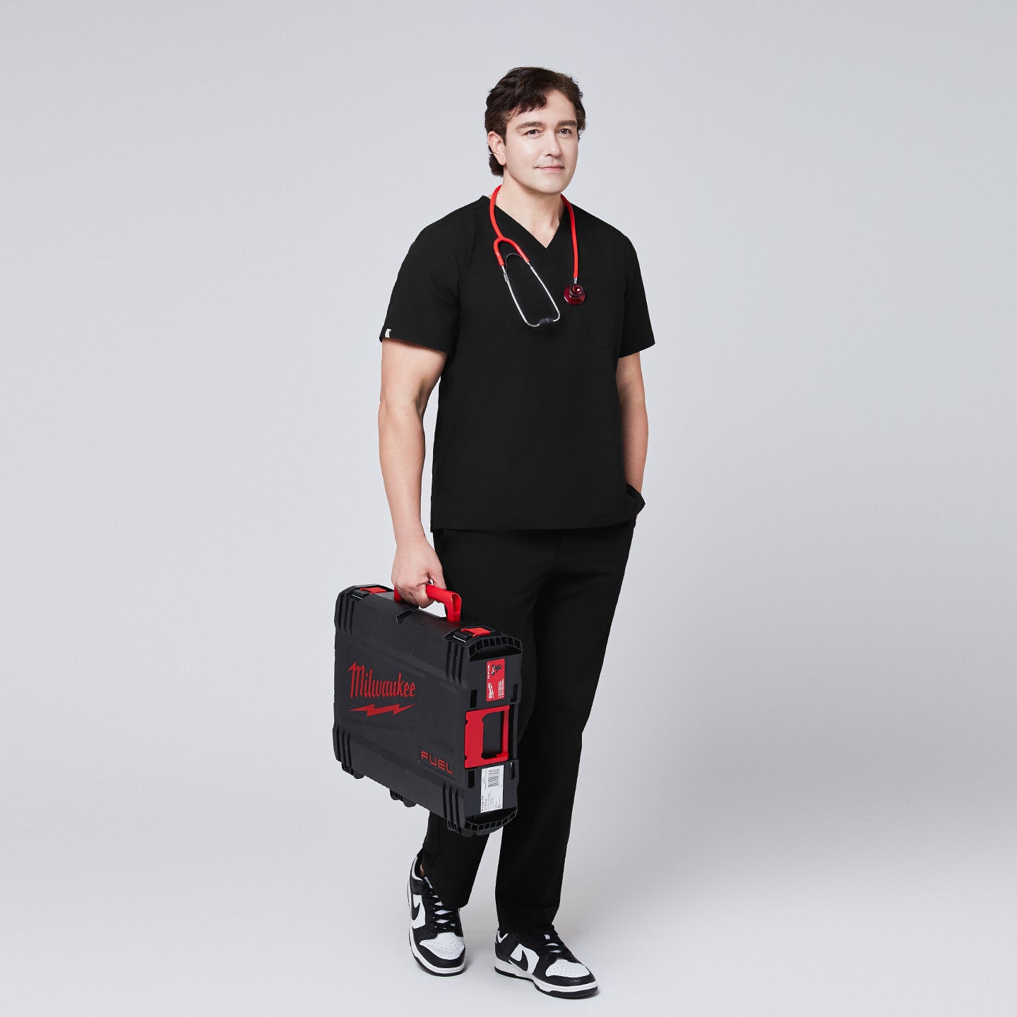 Image of Full-body view of a model wearing an eco black 3-pocket scrub top and matching pants, styled with a red stethoscope and carrying a Milwaukee tool case, paired with black sneakers,Eco Black