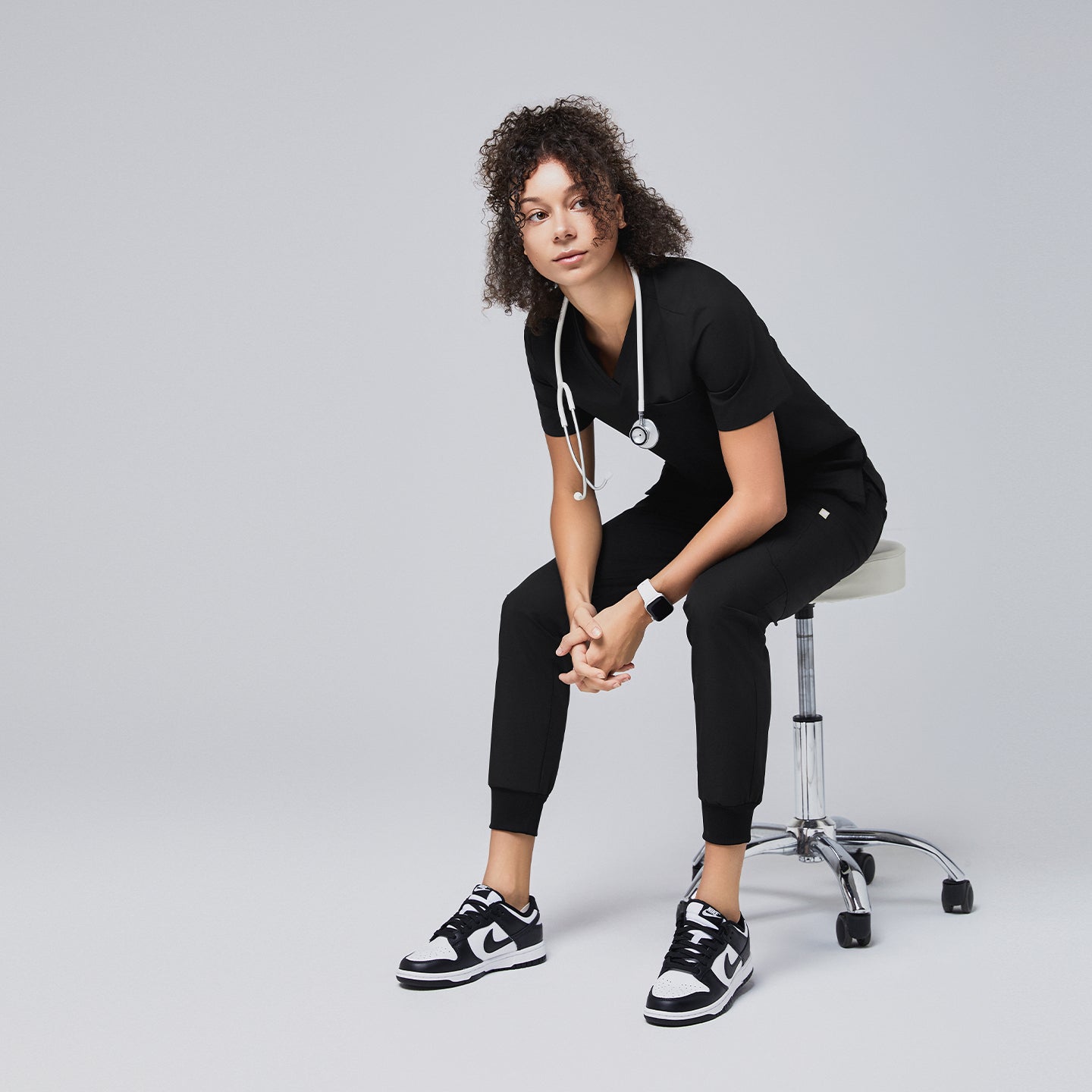 Woman wearing a black scrub set with a stethoscope, sitting on a stool in a relaxed pose,Eco Black