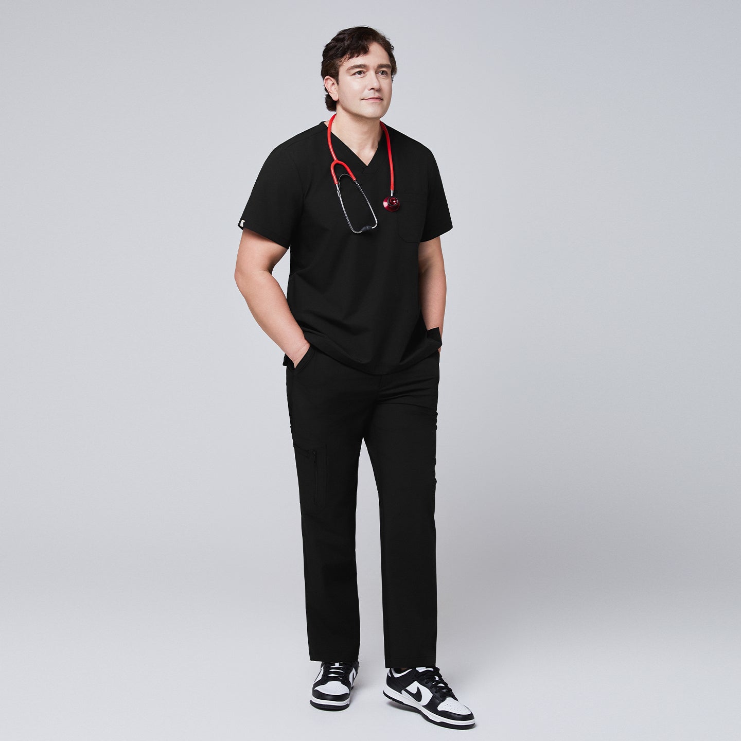 Image of Full-body view of a model wearing an eco black 3-pocket scrub top and matching pants, styled with a red stethoscope and black sneakers,Eco Black