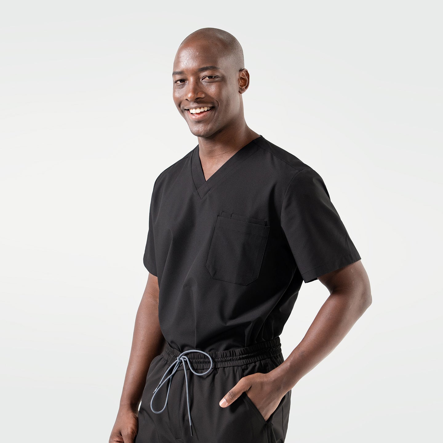The male model smiles while wearing an Eco Black scrub top with matching scrub pants, standing casually with one hand in his pocket.