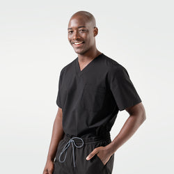 Image of The male model smiles while wearing an Eco Black scrub top with matching scrub pants, standing casually with one hand in his pocket.