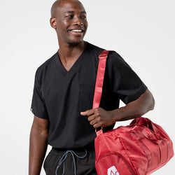 Image of Carrying a red duffel bag over his shoulder, the male model smiles while dressed in an Eco Black scrub top and matching pants.