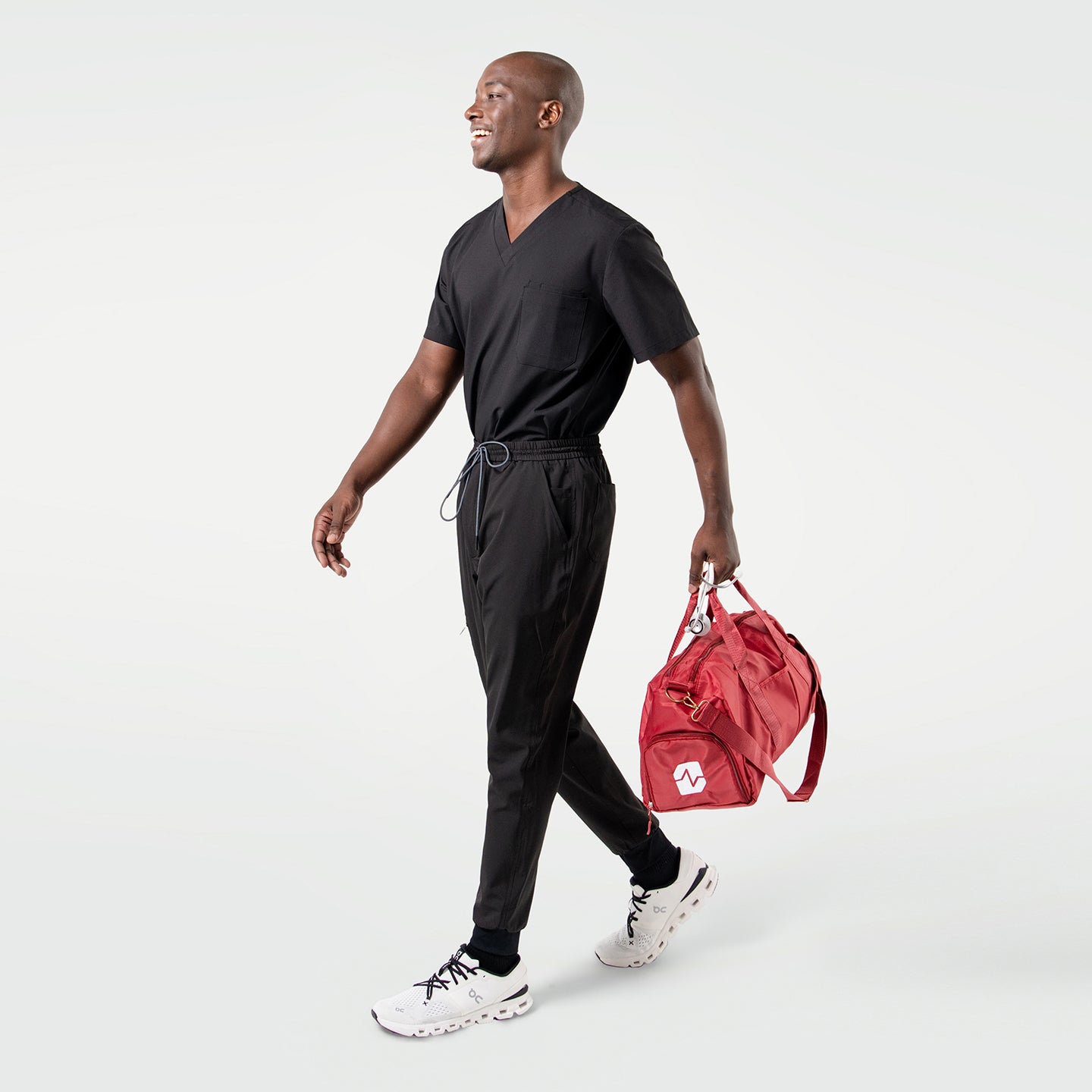 Image of Smiling model in an Eco Black 3 pocket scrub top with short sleeves and chest pocket, paired with matching scrub pants while carrying a red duffel bag.