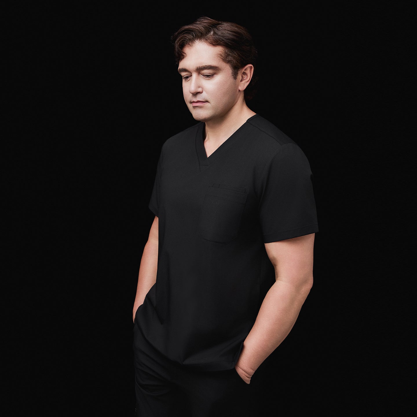Image of Side view of a model wearing an eco black 3-pocket scrub top with a V-neck design, hands in pockets, styled with matching scrub pants, against a black background,Eco Black