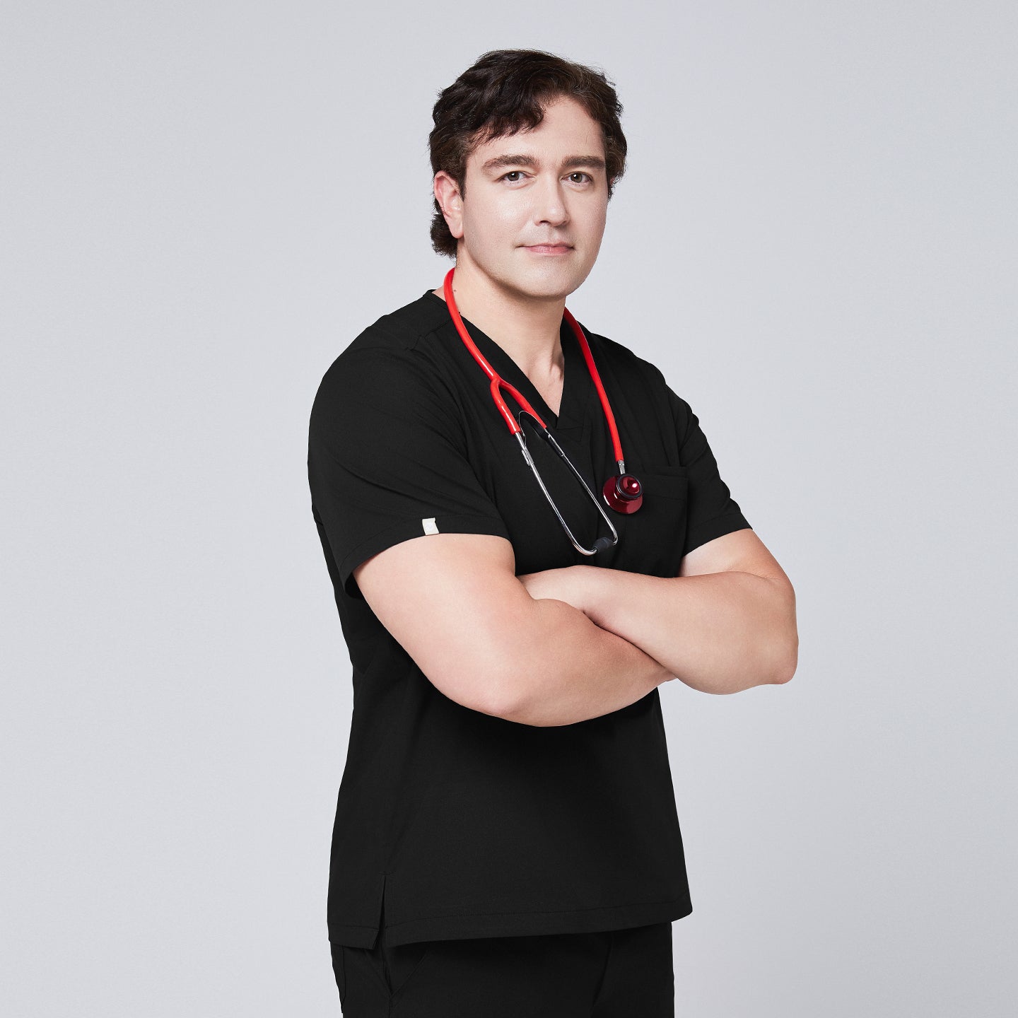 Image of Model wearing an eco black 3-pocket scrub top with a red stethoscope, arms crossed, styled with matching scrub pants,Eco Black