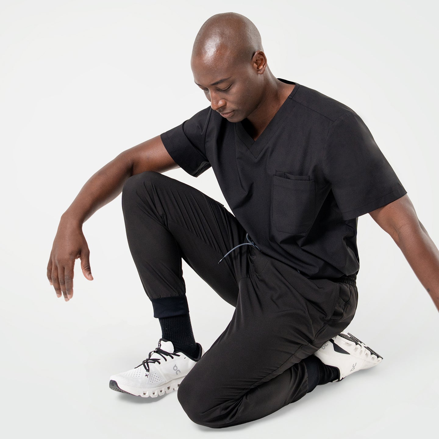 Kneeling pose in an Eco Black 3 pocket scrub top with short sleeves and chest pocket, paired with matching jogger scrub pants and white sneakers.