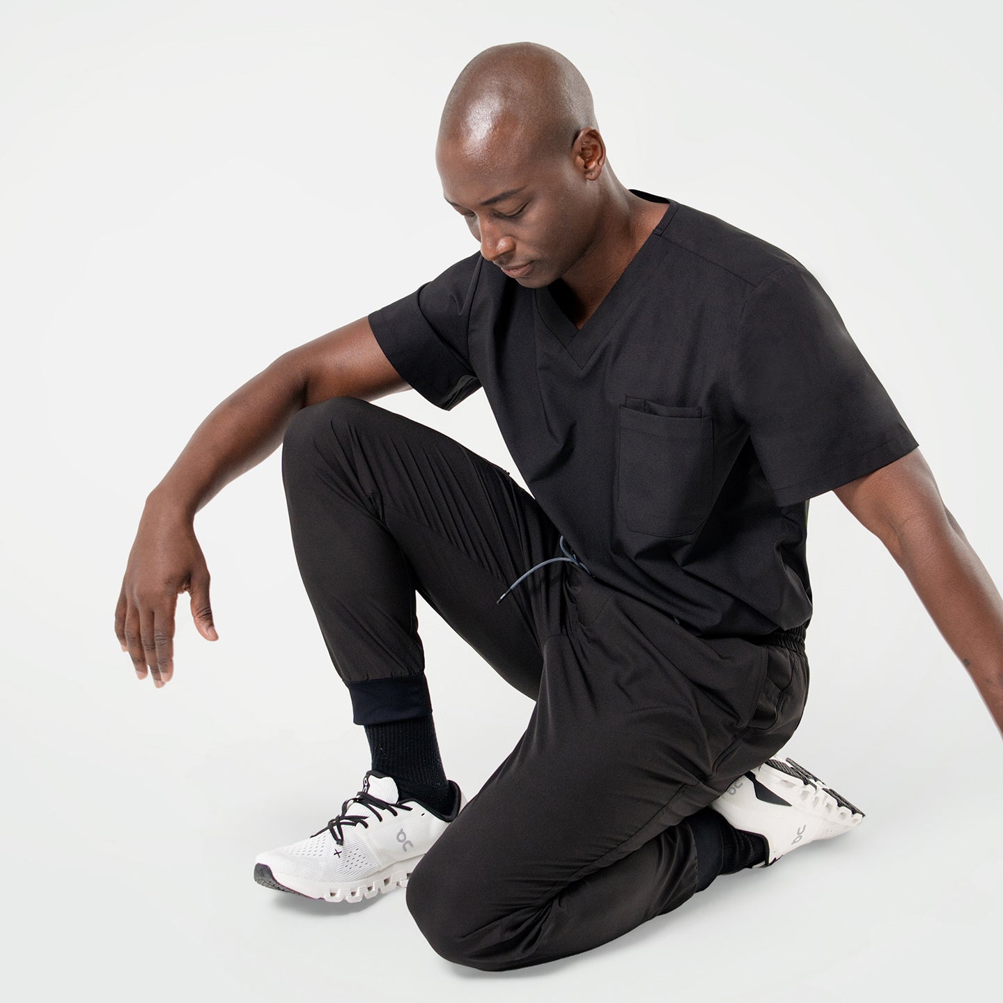 Image of Kneeling pose in an Eco Black 3 pocket scrub top with short sleeves and chest pocket, paired with matching jogger scrub pants and white sneakers.