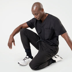 Image of Kneeling pose in an Eco Black 3 pocket scrub top with short sleeves and chest pocket, paired with matching jogger scrub pants and white sneakers.