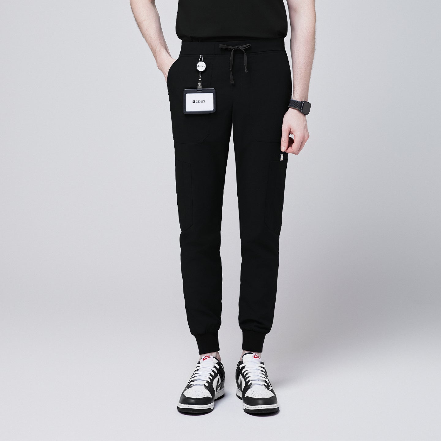 Image of Front view of black jogger scrub pants with 8 pockets, drawstring waistband, and cuffed ankles, paired with black and white sneakers and a name badge,Eco Black