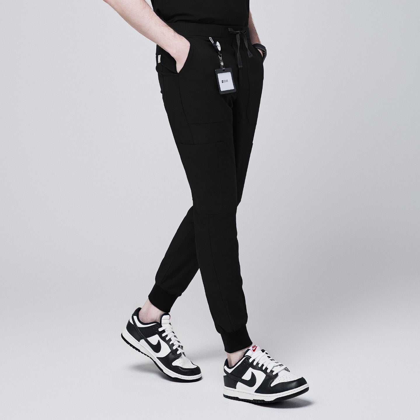 Image of Side angle of black jogger scrub pants with 8 pockets, drawstring waistband, and cuffed ankles, paired with black and white sneakers and a name badge,Eco Black