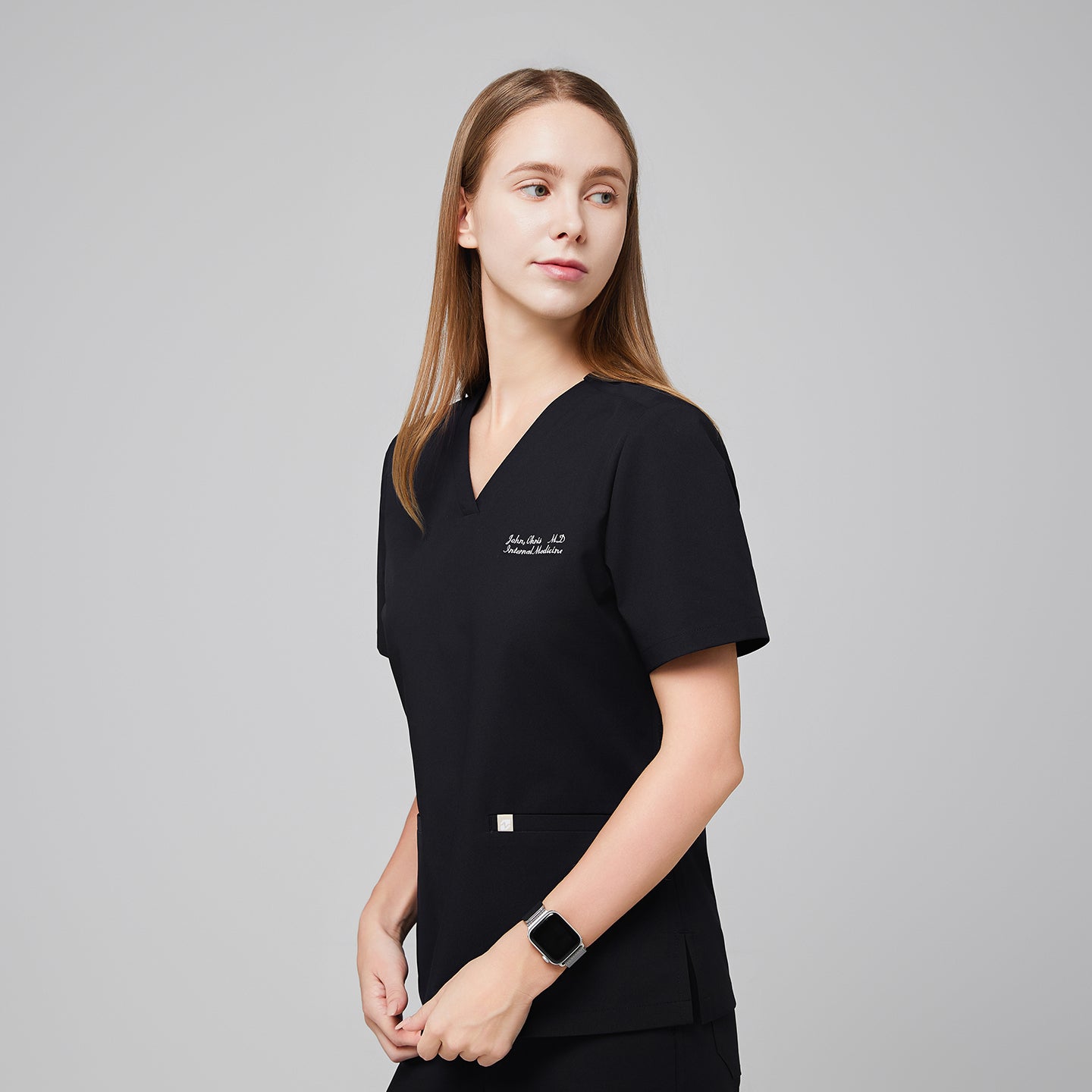 Woman wearing a black V-neck scrub top with embroidered name and side pockets, looking to the side,Eco Black