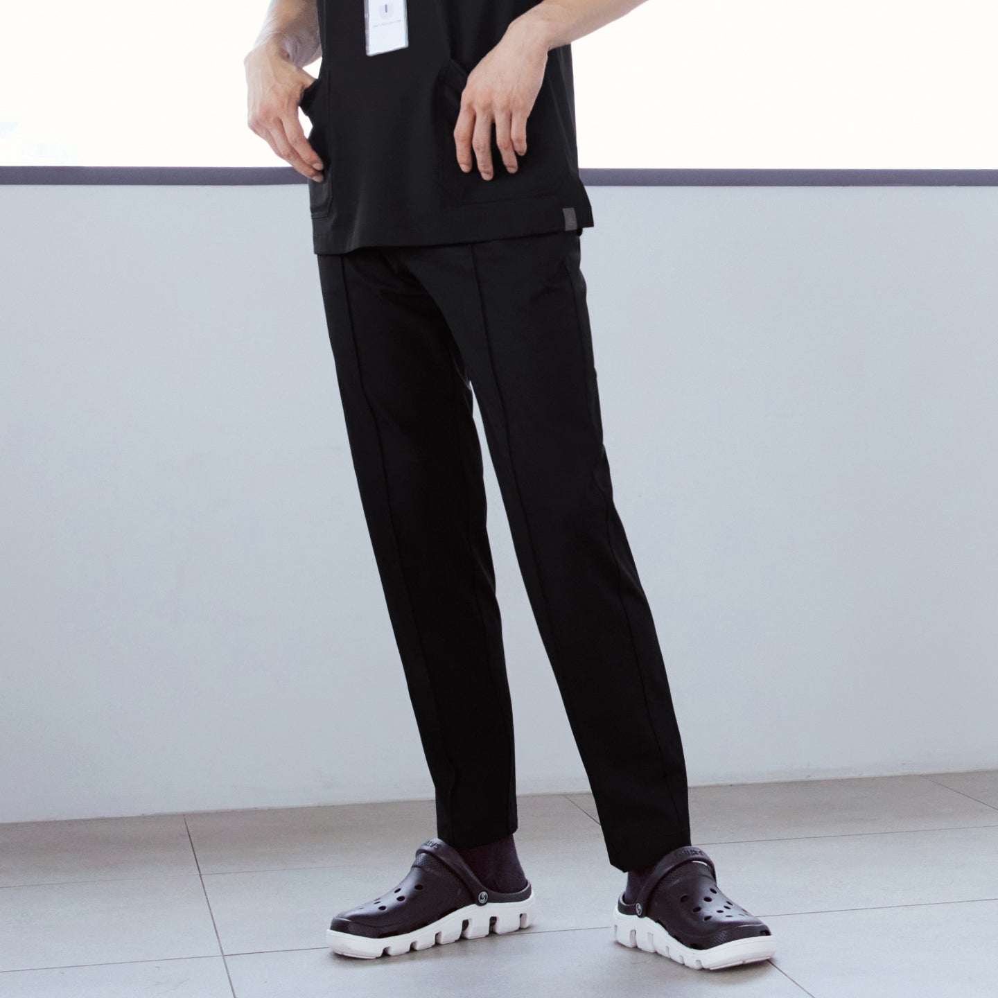 Image of Close-up of man wearing front zipper scrub top with chest and side pockets, paired with line banding scrub pants, hands in pockets, standing indoors,Eco Black
