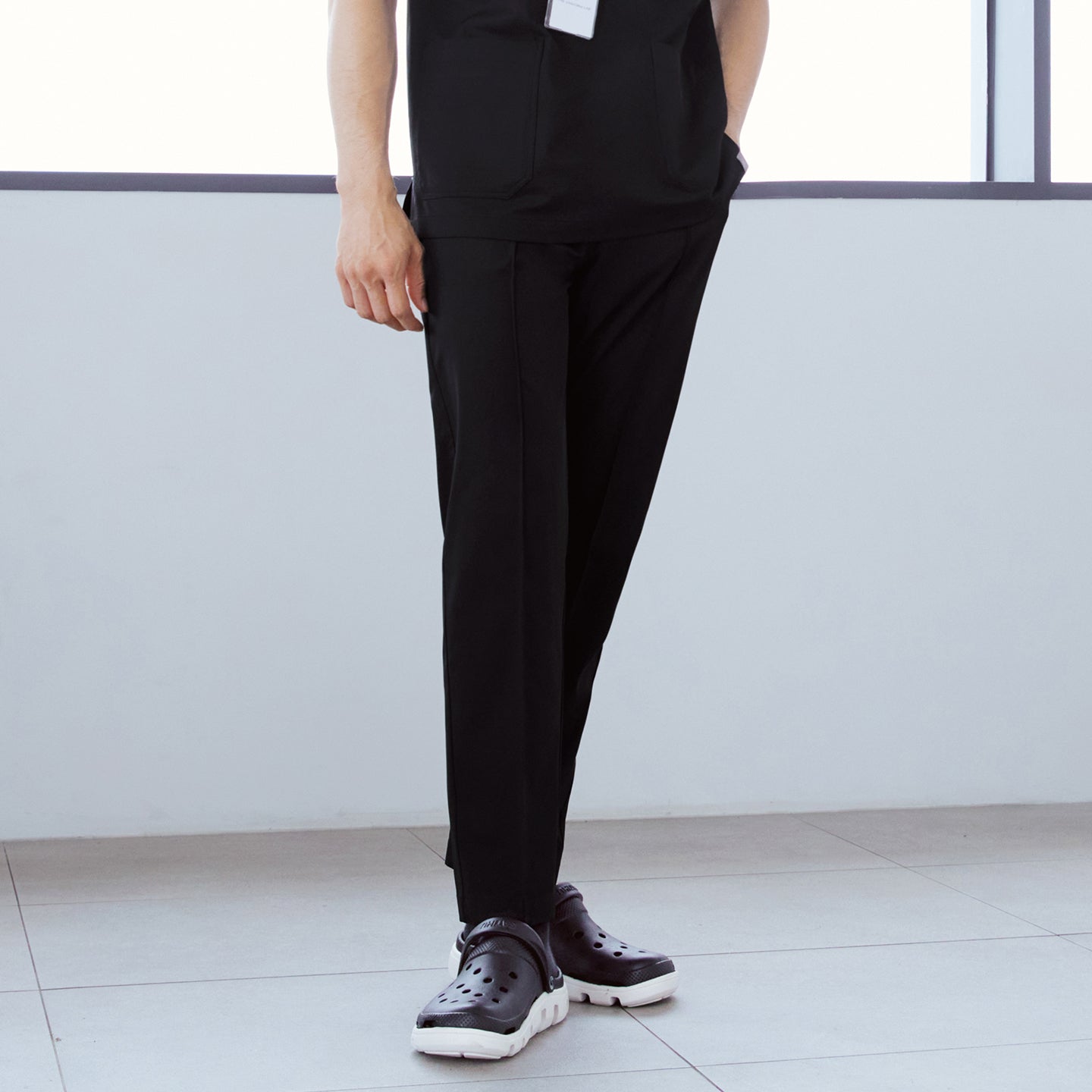 Close-up of man wearing front zipper scrub top with chest and side pockets, paired with banding scrub pants, standing indoors with one hand in pocket.,Eco Black