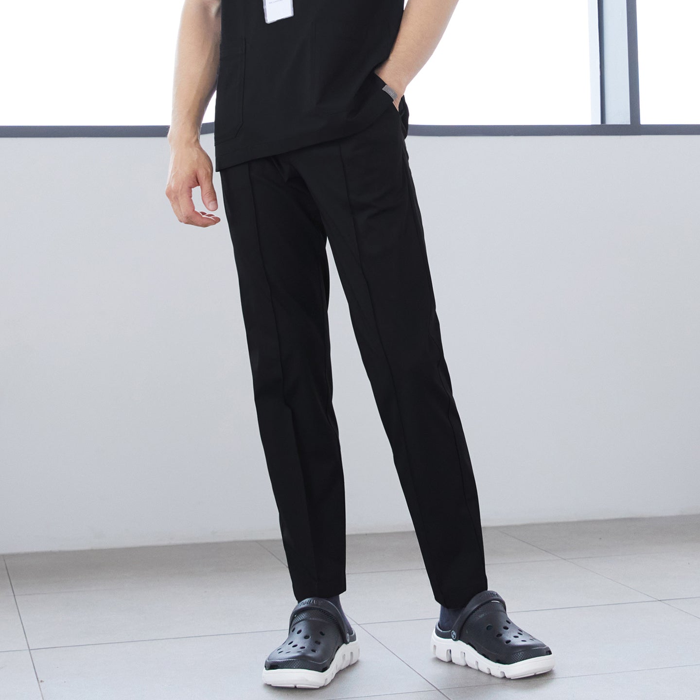Image of Close-up of man wearing front zipper scrub top with chest and side pockets, paired with straight-leg scrub pants, standing indoors with hands in pockets,Eco Black