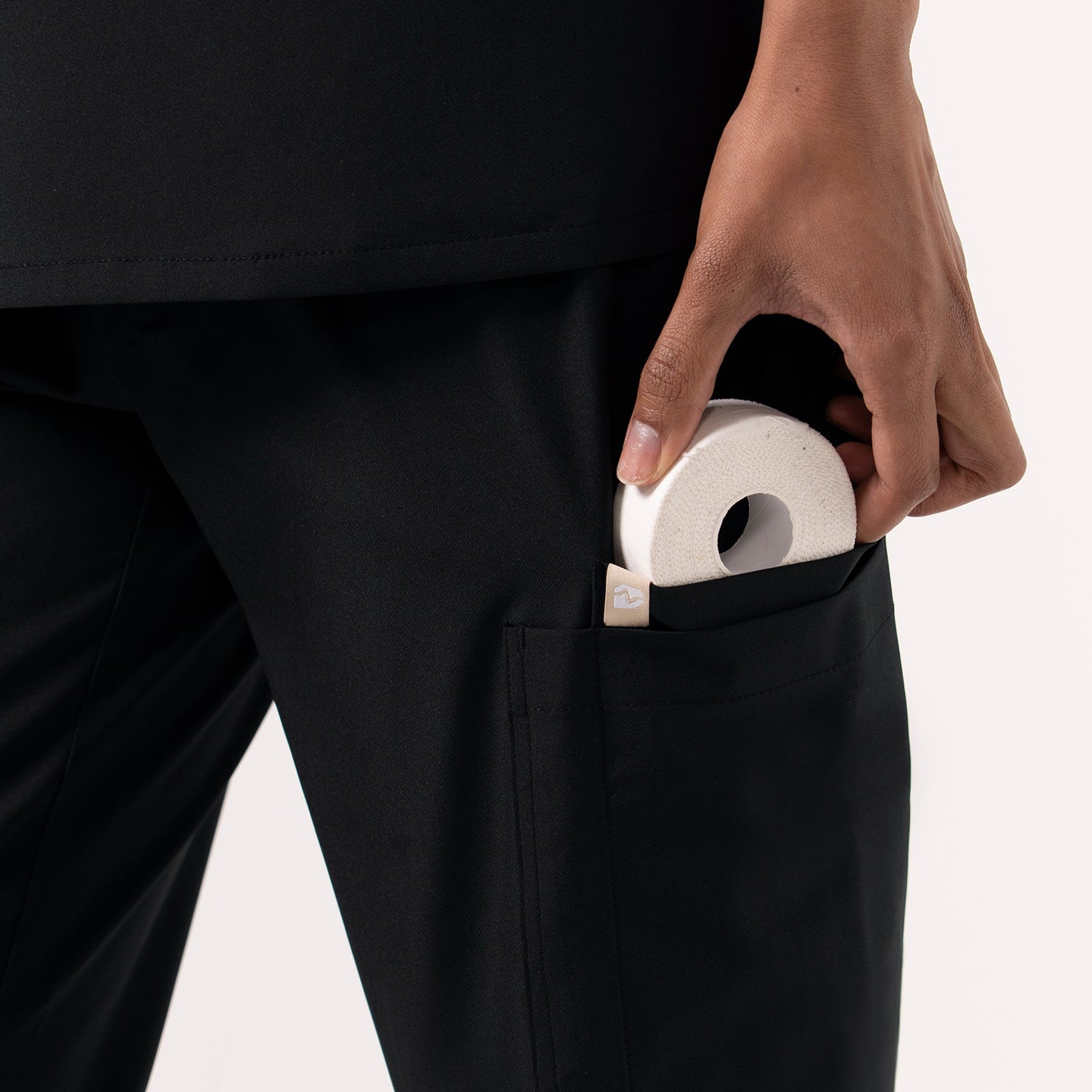 Image of Close-up view of a person wearing black jogger-style scrub pants, shown placing a small round medical item into the side cargo pocket. The fabric appears smooth and durable, and the pocket design is deep and functional, emphasizing storage convenience for healthcare professionals.