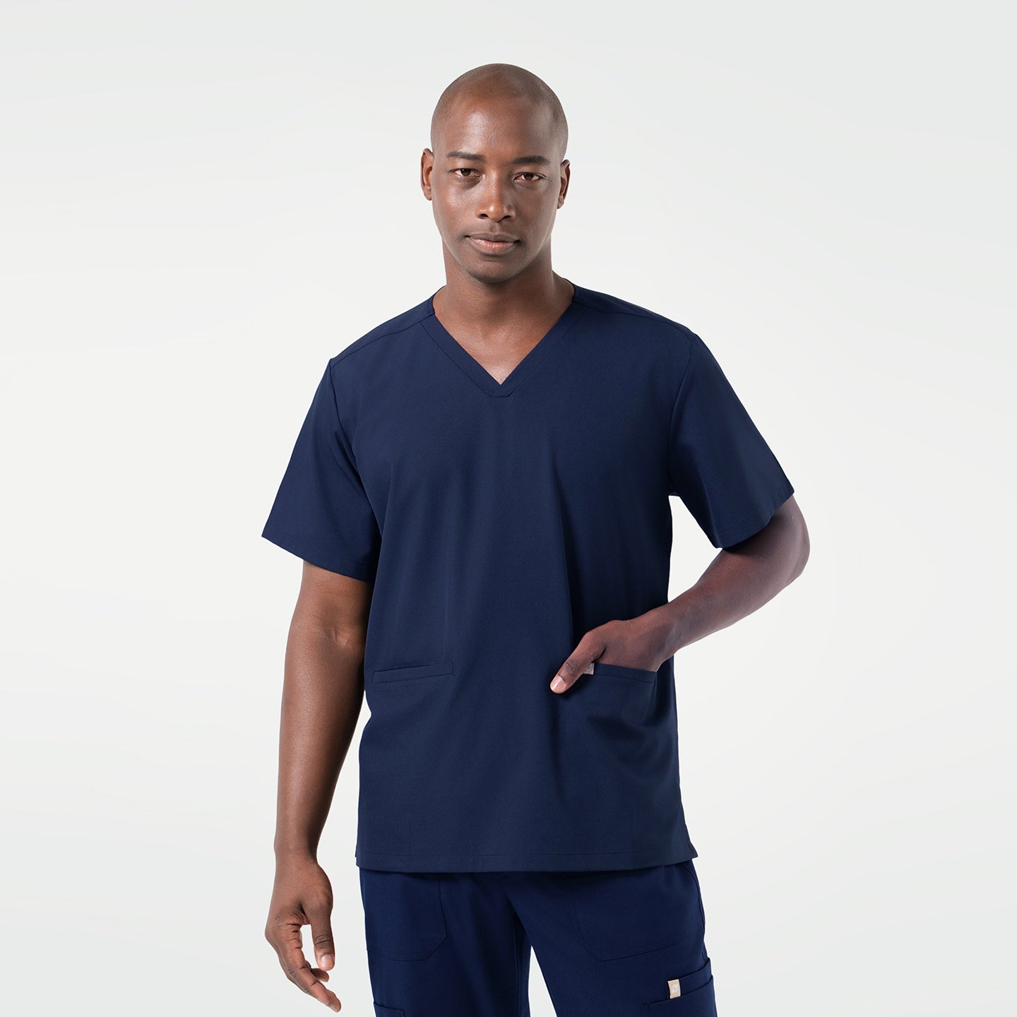 Wearing an Eco Navy scrub set, the male model stands facing forward with one hand placed in the front pocket, presenting a clean and professional look.