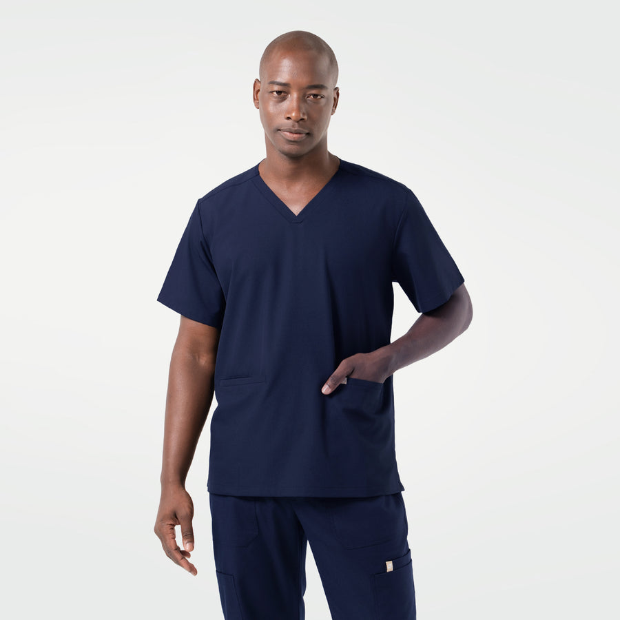 Wearing an Eco Navy scrub set, the male model stands facing forward with one hand placed in the front pocket, presenting a clean and professional look.