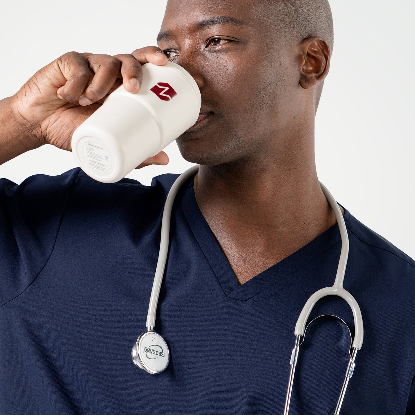 Dressed in an Eco Navy scrub top, the male model drinks from a white tumbler with a stethoscope resting around his neck.