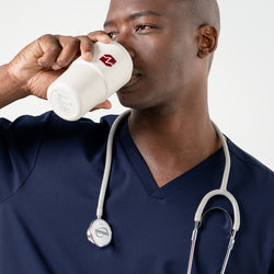 Image of Dressed in an Eco Navy scrub top, the male model drinks from a white tumbler with a stethoscope resting around his neck.