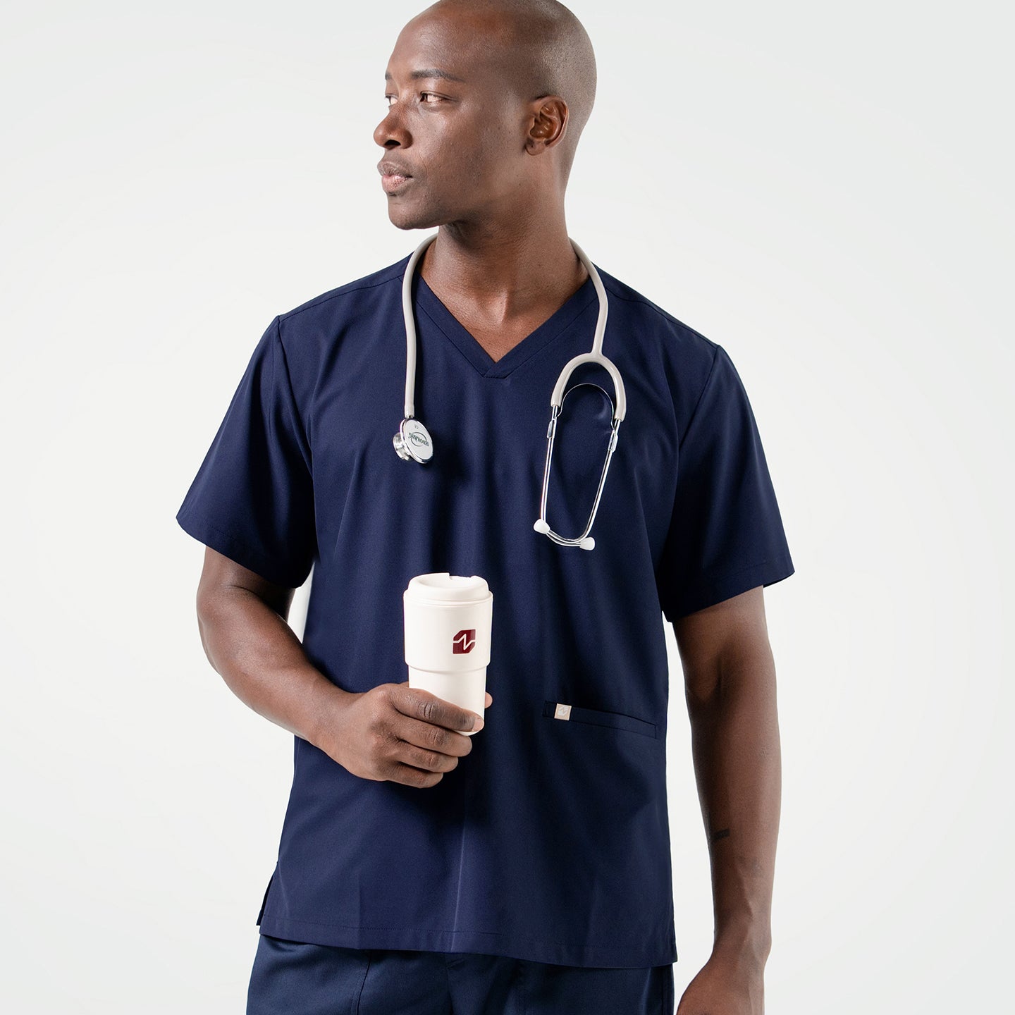 Holding a white tumbler in one hand, the male model wears an Eco Navy scrub set with a stethoscope around his neck while looking to the side.