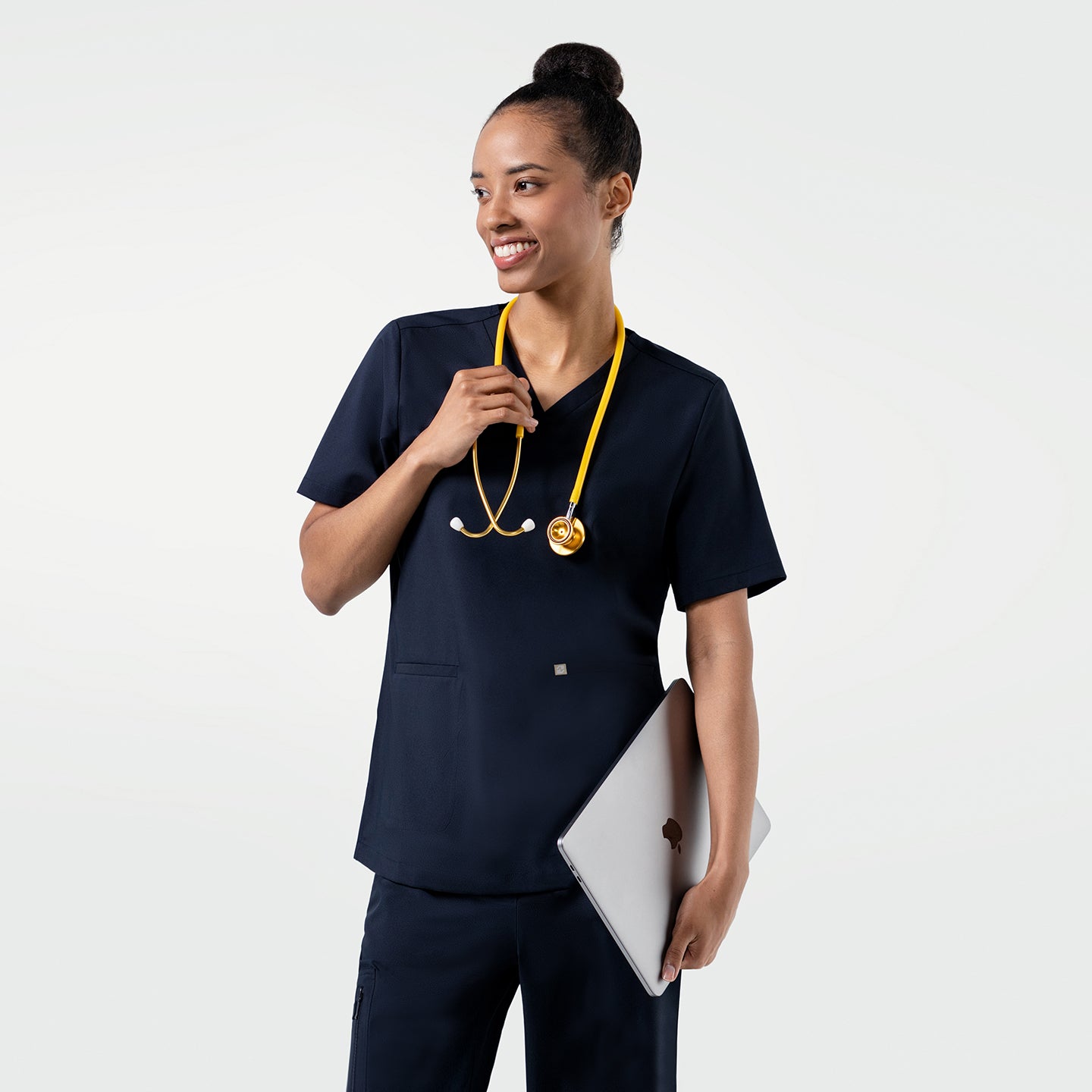 Model dressed in the Women's Eco Navy Double Pocket Scrub Top with pants, smiling with a yellow stethoscope and laptop in hand, upper body shot