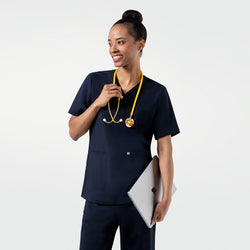 Image of Model dressed in the Women's Eco Navy Double Pocket Scrub Top with pants, smiling with a yellow stethoscope and laptop in hand, upper body shot