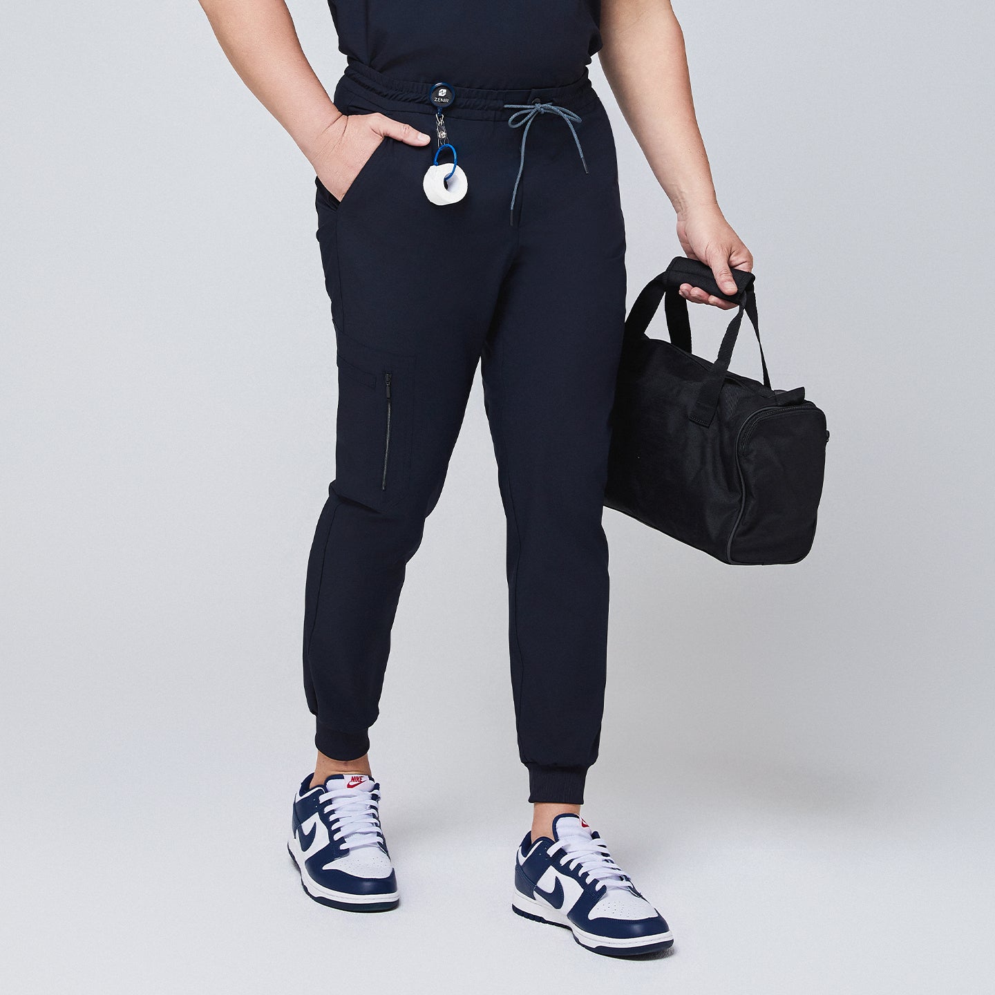 Image of Navy jogger scrub pants with a zipper pocket, drawstring waist, and attached badge clip, styled with a black bag,Eco Navy