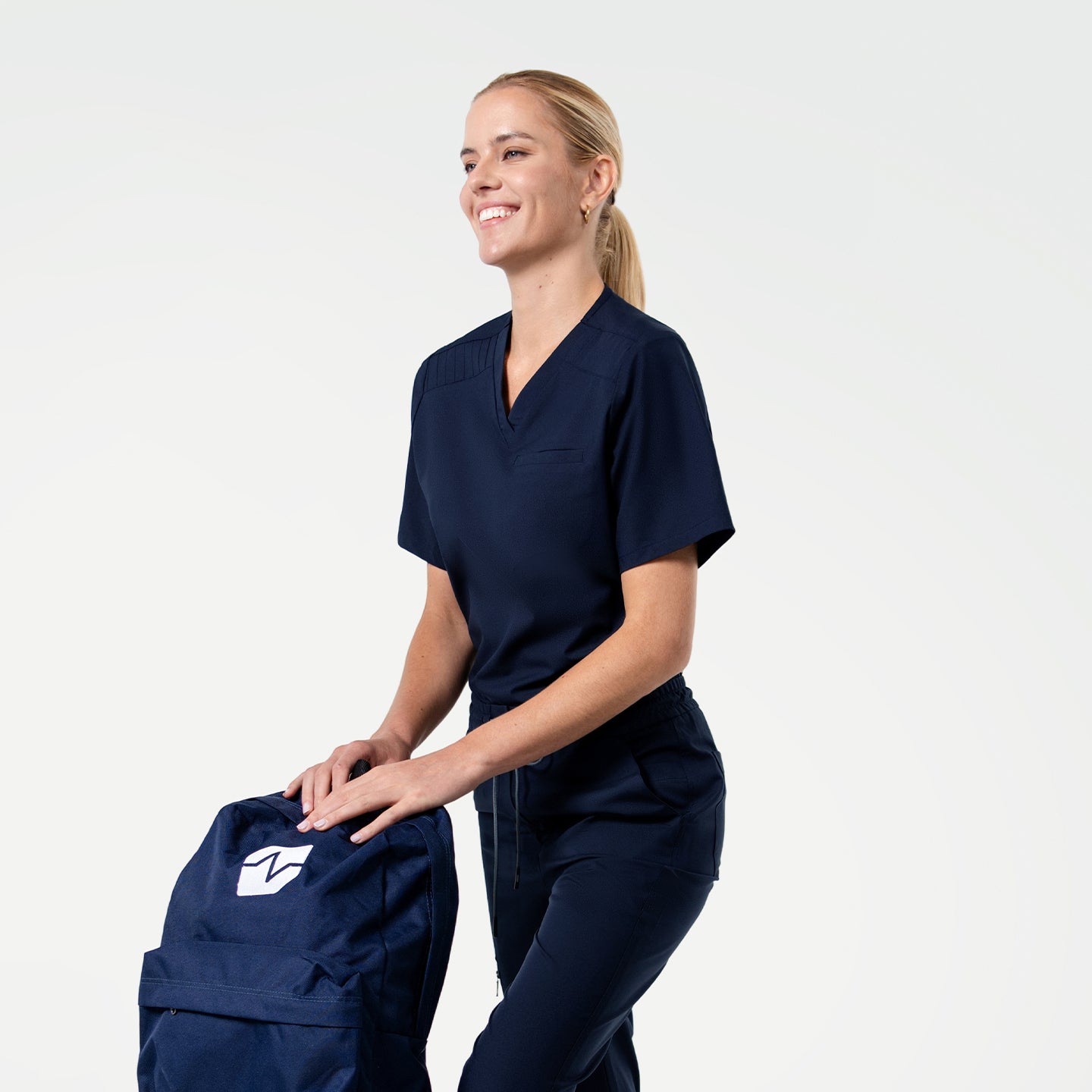 Image of Smiling in the Women's Eco Navy Pleated Shoulder Scrub Top and pants, holding a navy backpack, upper body view