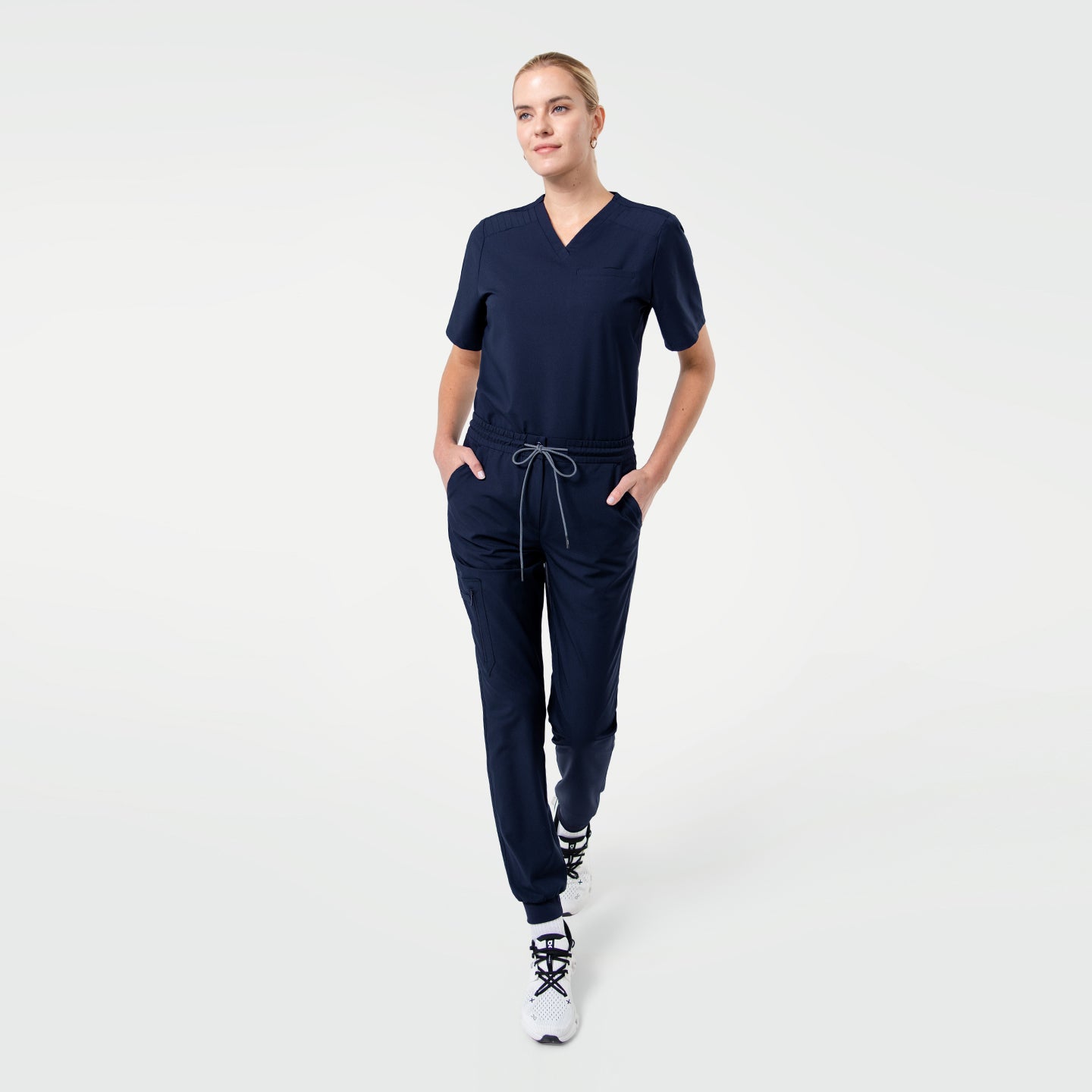 Image of Model walking in a navy V-neck pleated-shoulder scrub top and matching jogger scrub pants, hands in pockets, against a clean light background.