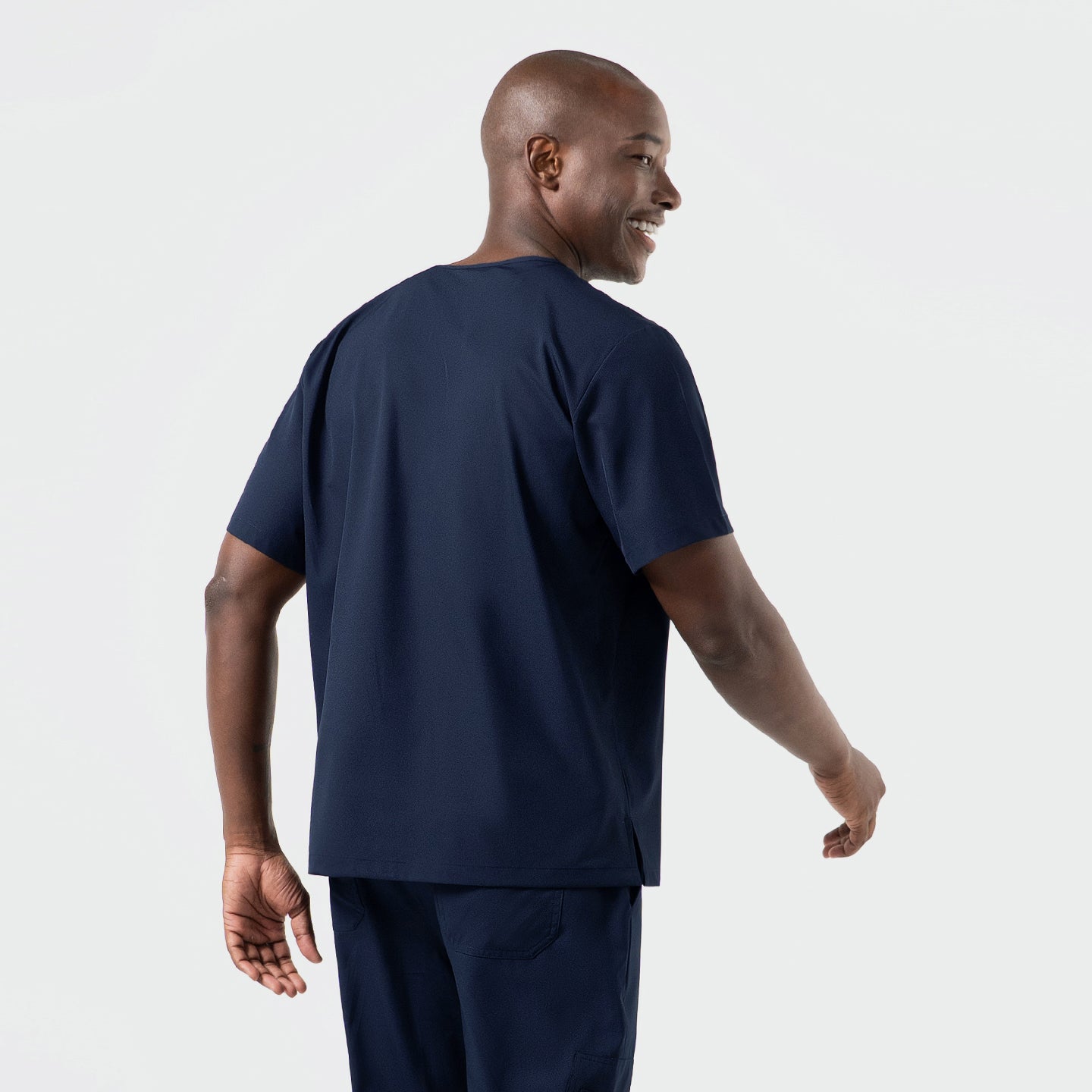 Image of Back and side-angle view of a man wearing an Eco-Navy single-pocket scrub top, highlighting the smooth performance fabric, relaxed short-sleeve fit, and clean, minimal silhouette paired with matching scrub pants.