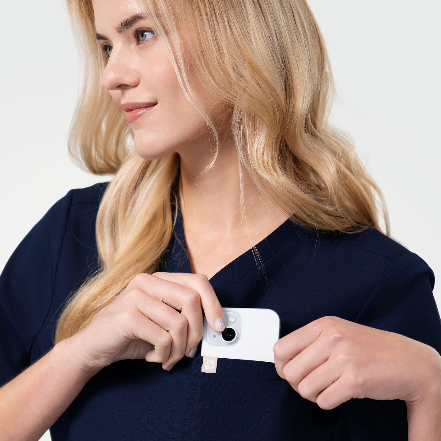 Image of A women’s Eco Navy single pocket scrub top is shown. The model places a small white device into the chest pocket, highlighting the storage feature.
