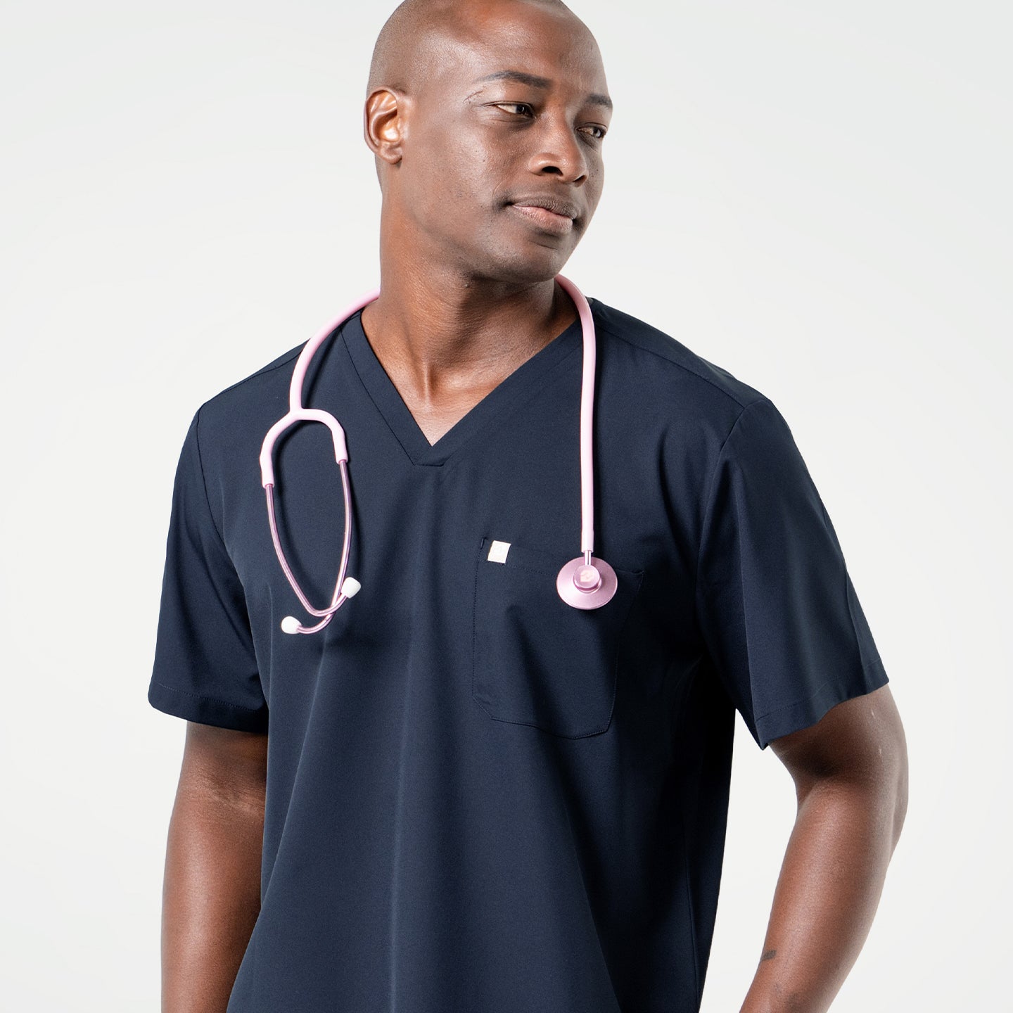 Wearing an Eco Navy scrub top, the male model looks to the side with a light pink stethoscope draped around his neck.