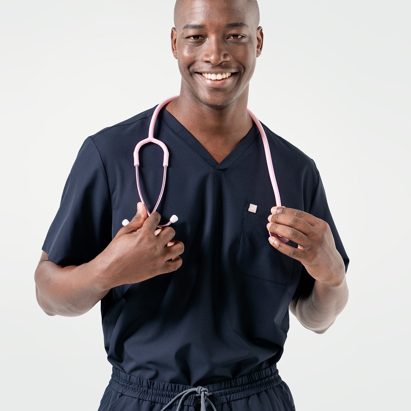 Smiling confidently, the male model wears an Eco Navy scrub top with a pink stethoscope draped around his neck.