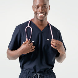 Image of Smiling confidently, the male model wears an Eco Navy scrub top with a pink stethoscope draped around his neck.
