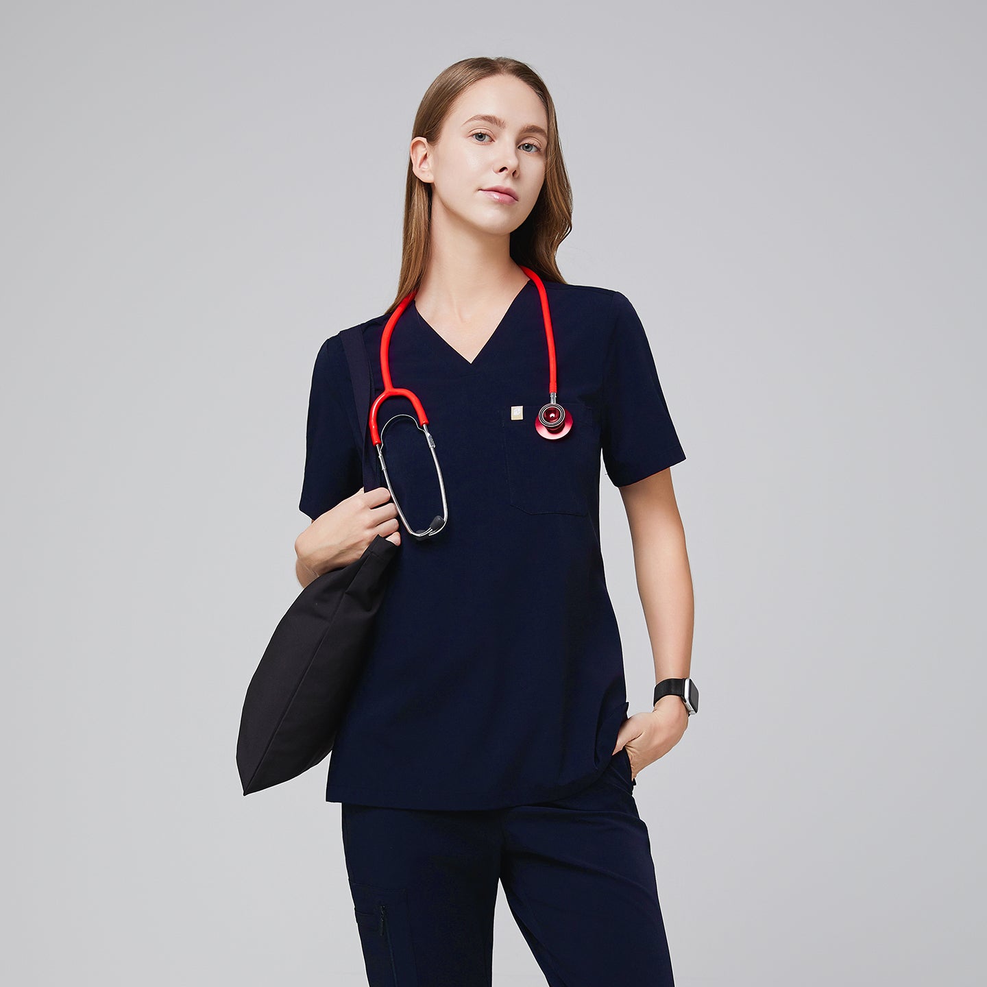Image of Women wearing navy scrub top with single pocket paired with matching scrub pants, with a red stethoscope around her neck and pillow by her side,Eco navy