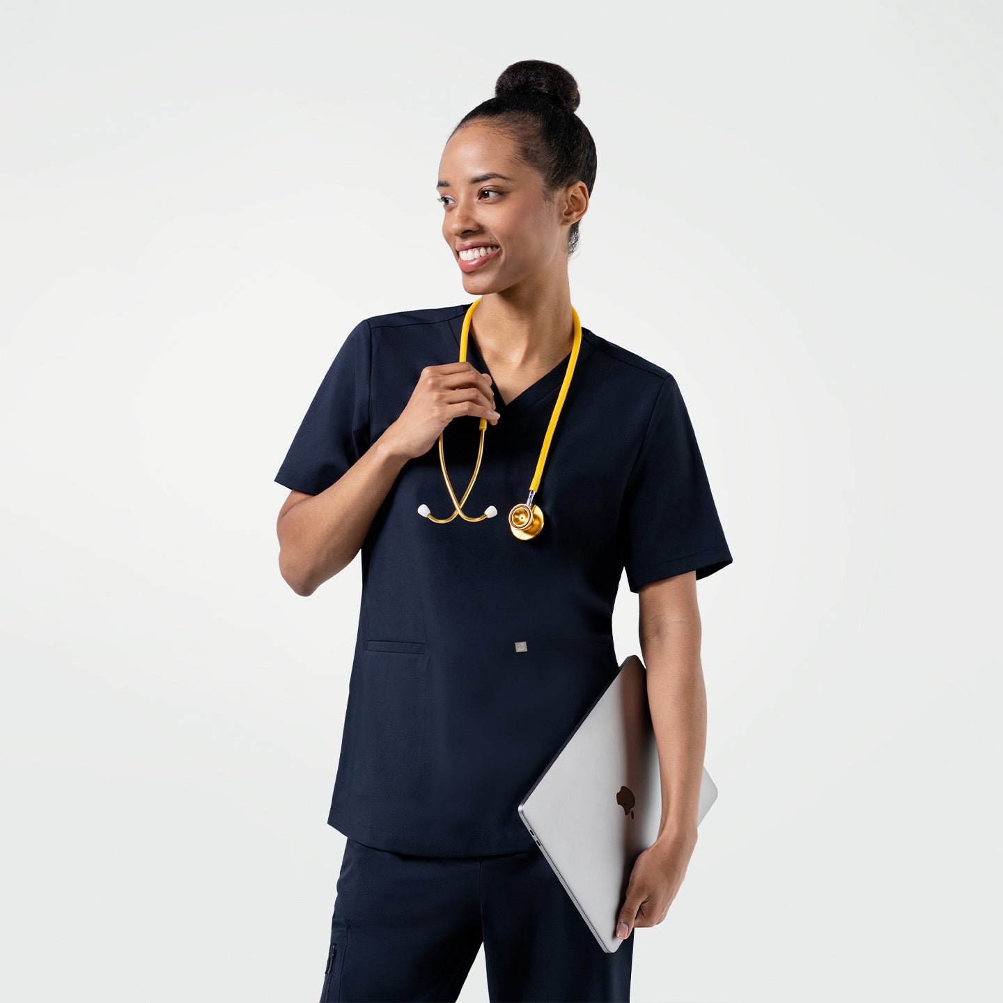Model dressed in the Women's Eco Navy Single Pocket Scrub Top with pants, smiling with a yellow stethoscope and laptop in hand, upper body shot