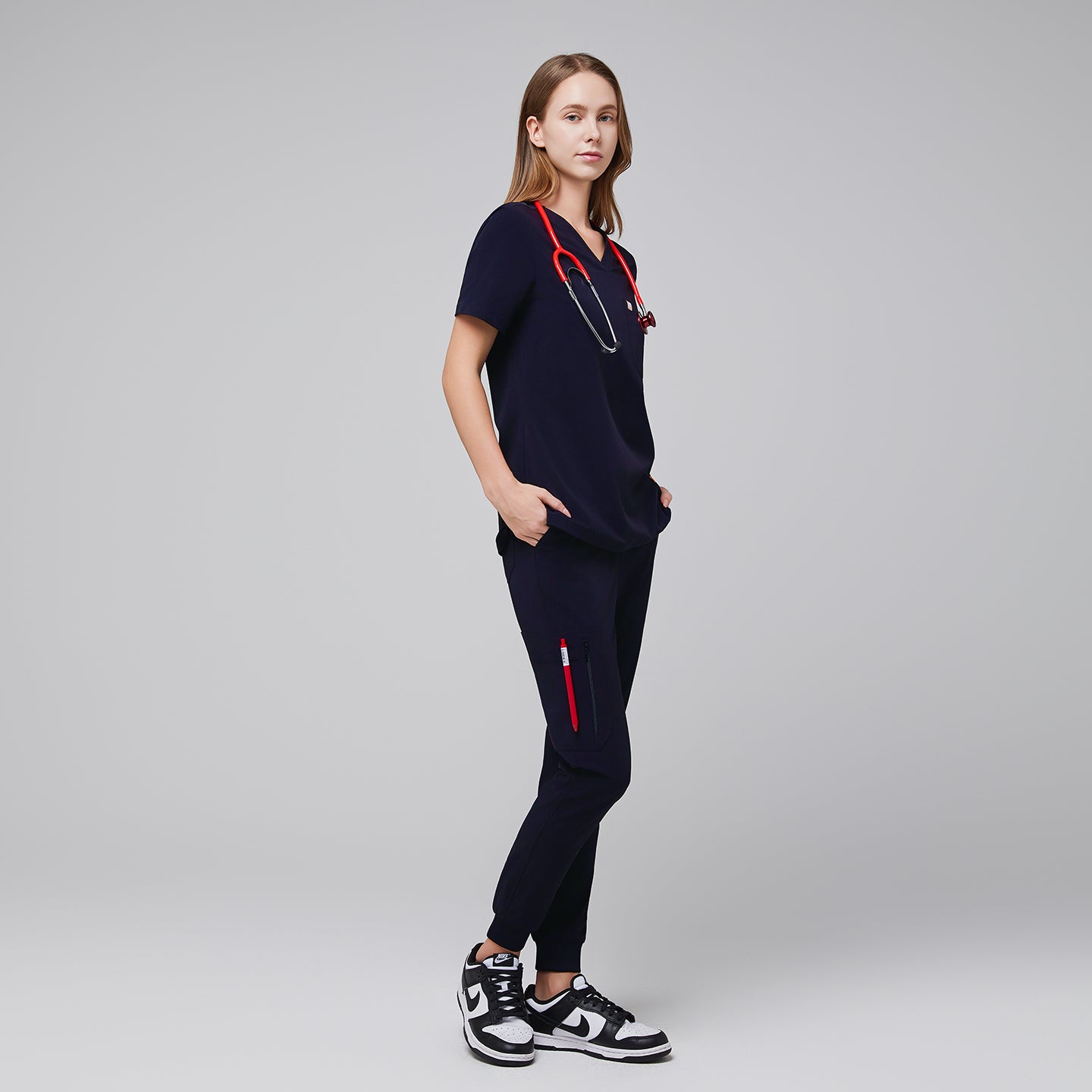 Image of Navy scrub top with single pocket paired with matching scrub pants, with a red stethoscope around their neck and pen in pants pocket and black sneakers,Eco navy